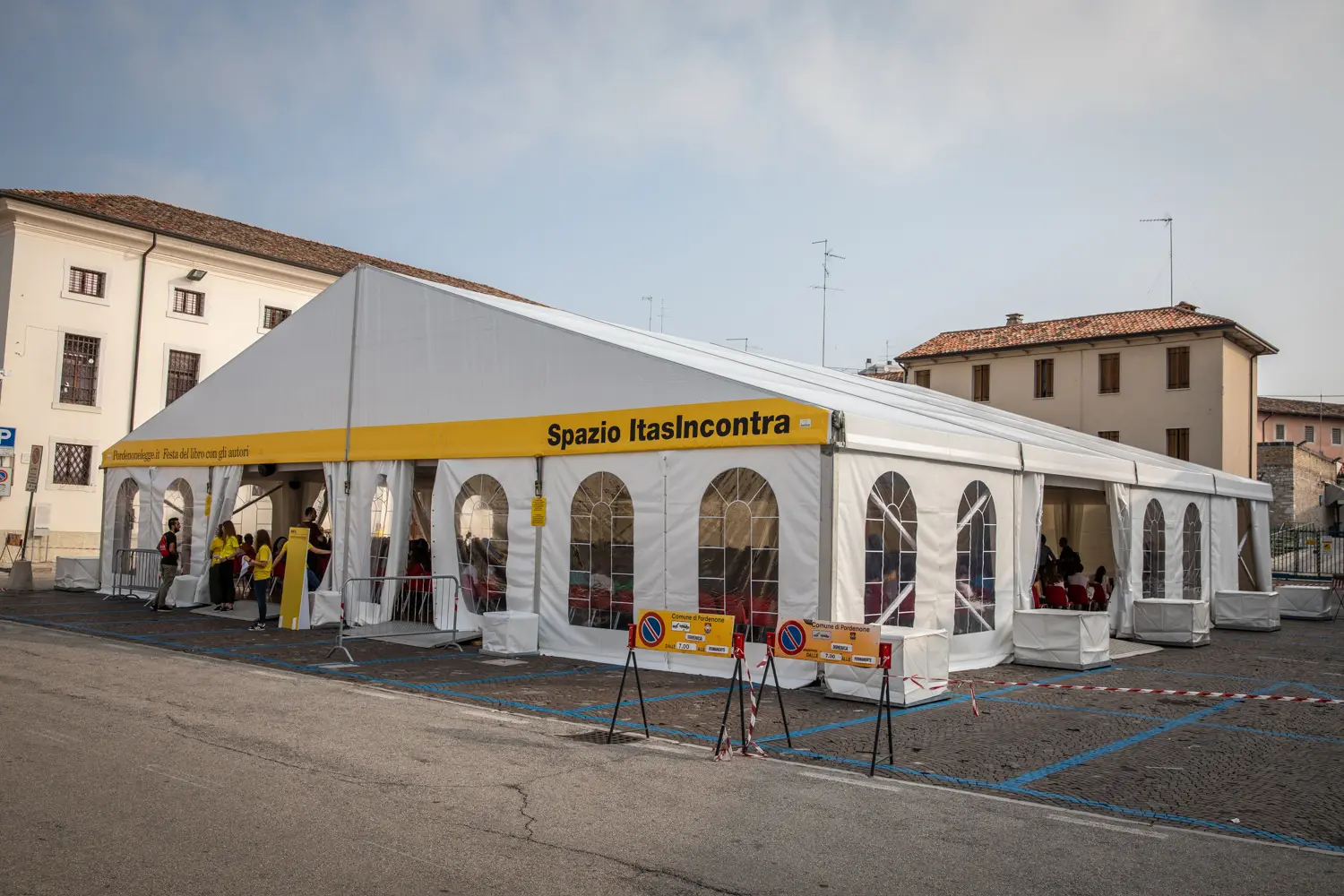 Una grande tenda bianca con finestre di plastica ad arco, con la scritta "Spazio ItasIncontra", è stata installata in un'area pavimentata. Cartelli e una barriera di corda segnano l'ingresso. Le persone sono radunate all'interno e gli edifici vicini sono visibili sotto un cielo parzialmente nuvoloso.