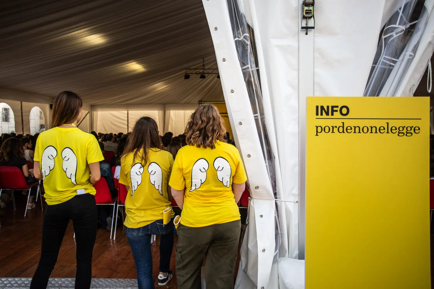 Tre persone con magliette gialle con un fantasma bianco disegnato sul retro sono in piedi vicino a un cartello informativo giallo con la scritta "pordenonelegge". Si trovano all'ingresso di una tenda piena di partecipanti seduti.