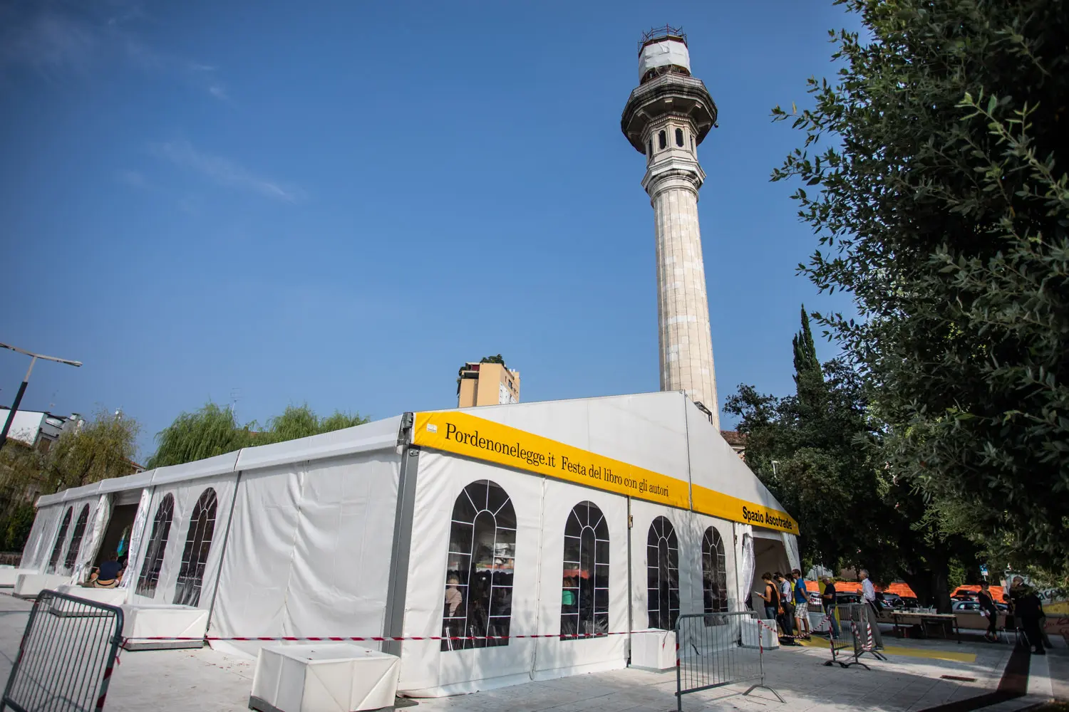 Un grande tendone bianco per eventi con persone all'interno è allestito accanto a una torre alta e slanciata contro un cielo azzurro terso. Il tendone ha uno striscione giallo con del testo. Intorno al tendone sono visibili alcuni alberi e barriere.