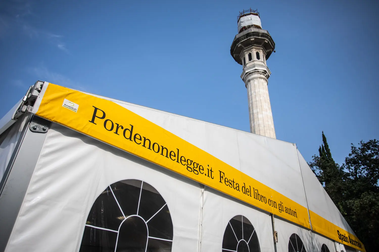 Una tenda bianca con finestre ad arco espone uno striscione giallo con la scritta "Pordenonelegge.it Festa del libro con gli autori". Sullo sfondo, contro il cielo azzurro, è parzialmente visibile una torre alta e decorata.
