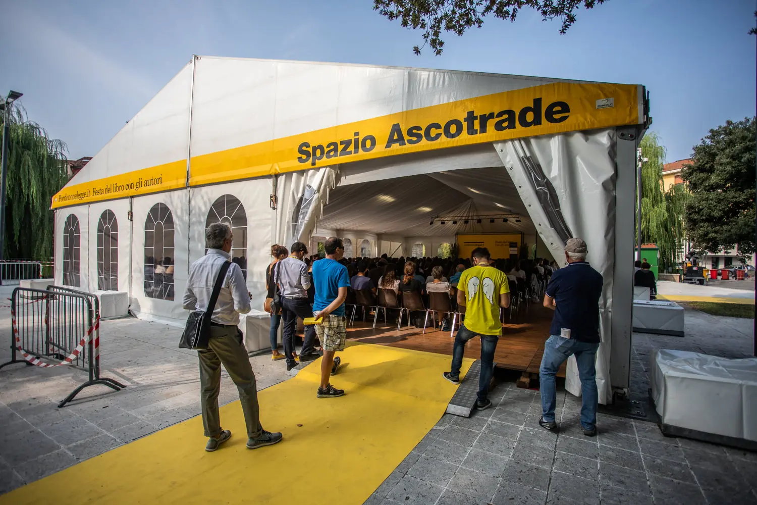Persone che entrano in una grande tenda bianca con la scritta "Spazio Ascotrade" e un cartello giallo. Un tappeto giallo conduce all'interno. La tenda è allestita all'aperto, con alberi e una folla leggera radunata all'ingresso.