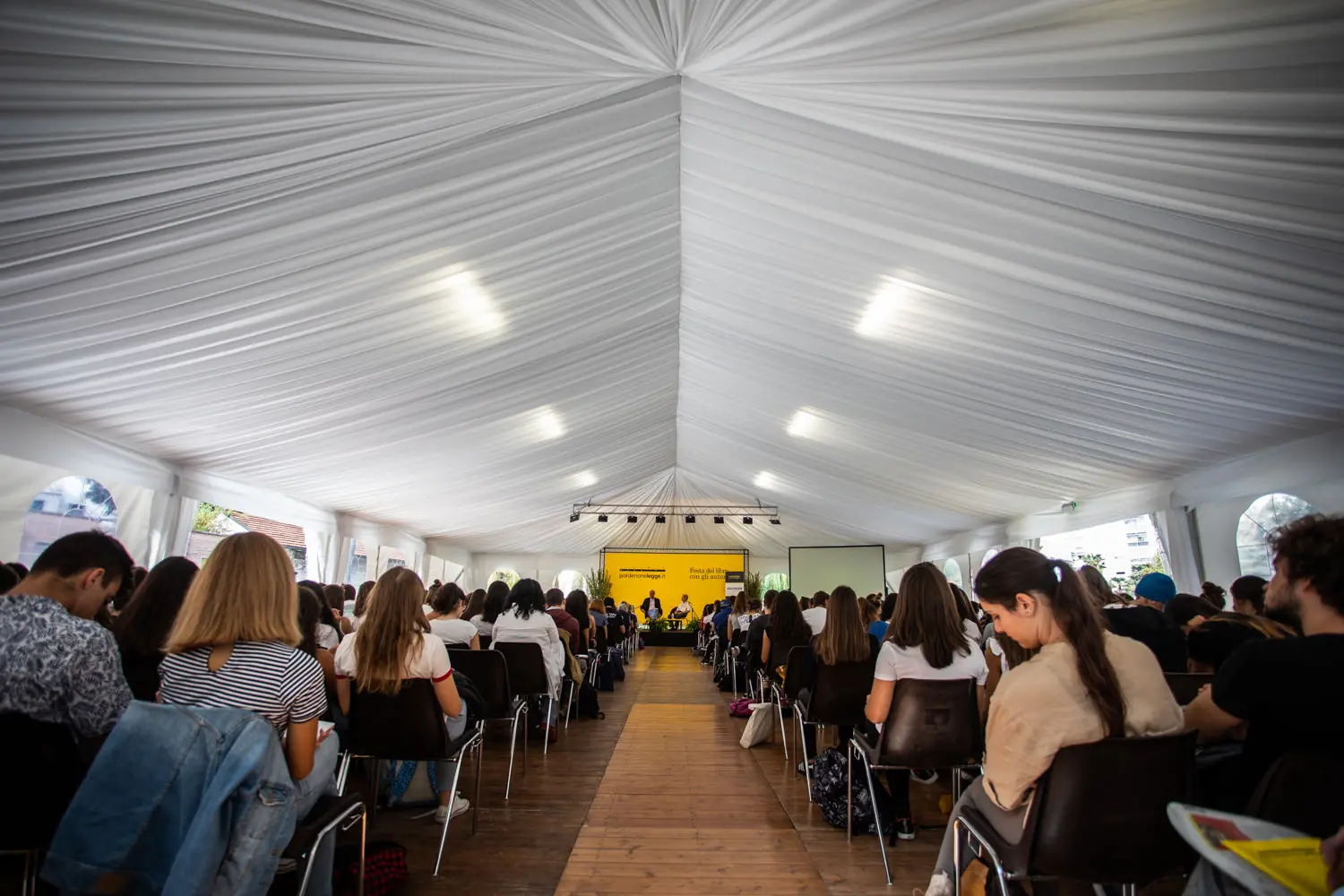 Un folto gruppo di persone sedute in fila sotto una tenda bianca, di fronte a un palco con uno sfondo giallo. L'ambientazione sembra quella di una conferenza o di un seminario formale. L'illuminazione è soffusa e l'atmosfera è concentrata e attenta.