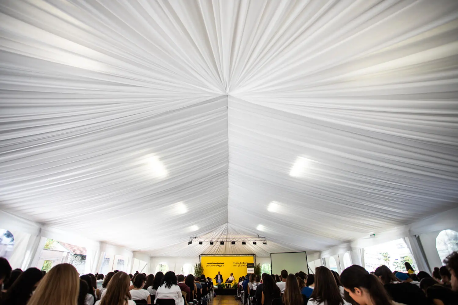 Un grande raduno si tiene all'interno di una tenda bianca con un soffitto drappeggiato. I partecipanti sono seduti di fronte a un palco con uno sfondo giallo e degli altoparlanti. L'atmosfera è formale e concentrata.