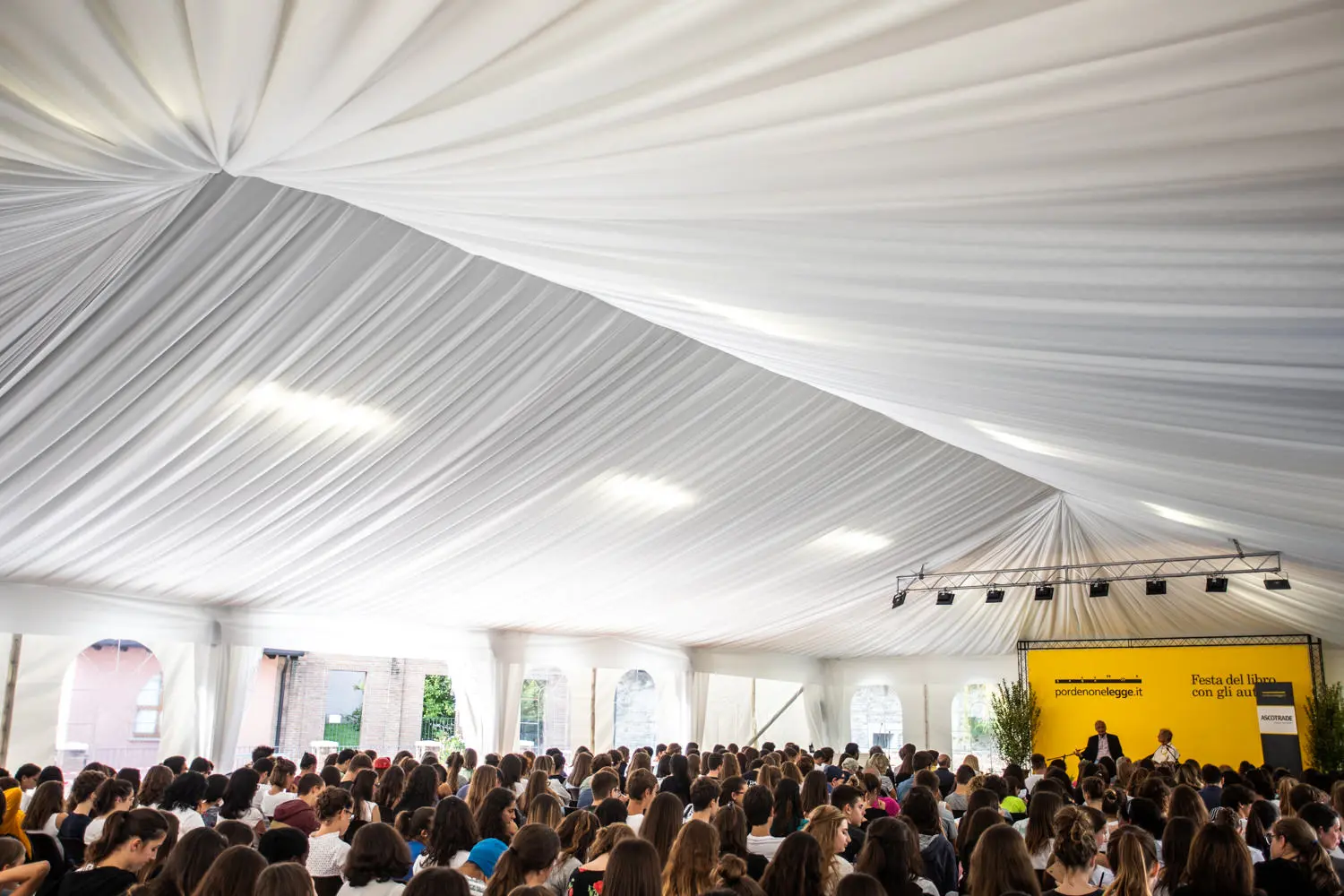 Un folto gruppo di persone è seduto sotto una spaziosa tenda bianca per eventi. Davanti, un oratore è in piedi vicino a uno sfondo giallo con del testo, e si rivolge al pubblico. Il soffitto della tenda è drappeggiato con un tessuto bianco fluente.