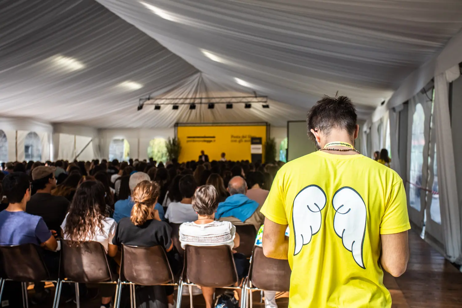 Un folto gruppo di persone sedute in un tendone ascolta un oratore sul palco. In primo piano, una persona che indossa una maglietta gialla brillante con ali d'angelo bianche sulla schiena è in piedi di fronte al pubblico.