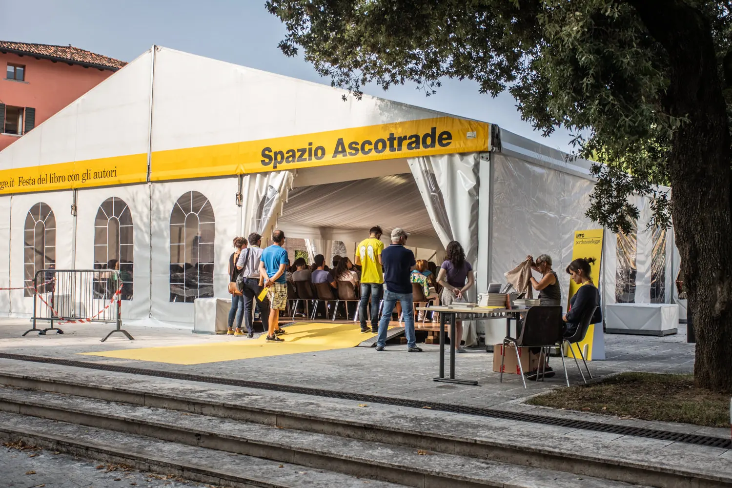 Un tendone bianco per eventi con un cartello giallo con la scritta "Spazio Ascotrade". Le persone entrano e si radunano attorno all'ingresso. Nelle vicinanze, personale in camice gialle fornisce assistenza. L'ambientazione è all'aperto con un grande albero in primo piano.