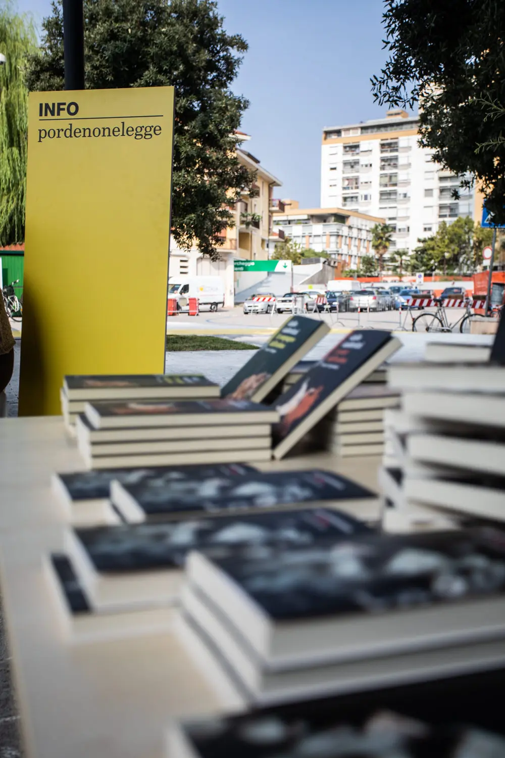 Un tavolo con pile di libri a fuoco, con un cartello giallo "INFO pordenonelegge" di lato. Sullo sfondo, sono visibili un albero, edifici e un cantiere con barriere.