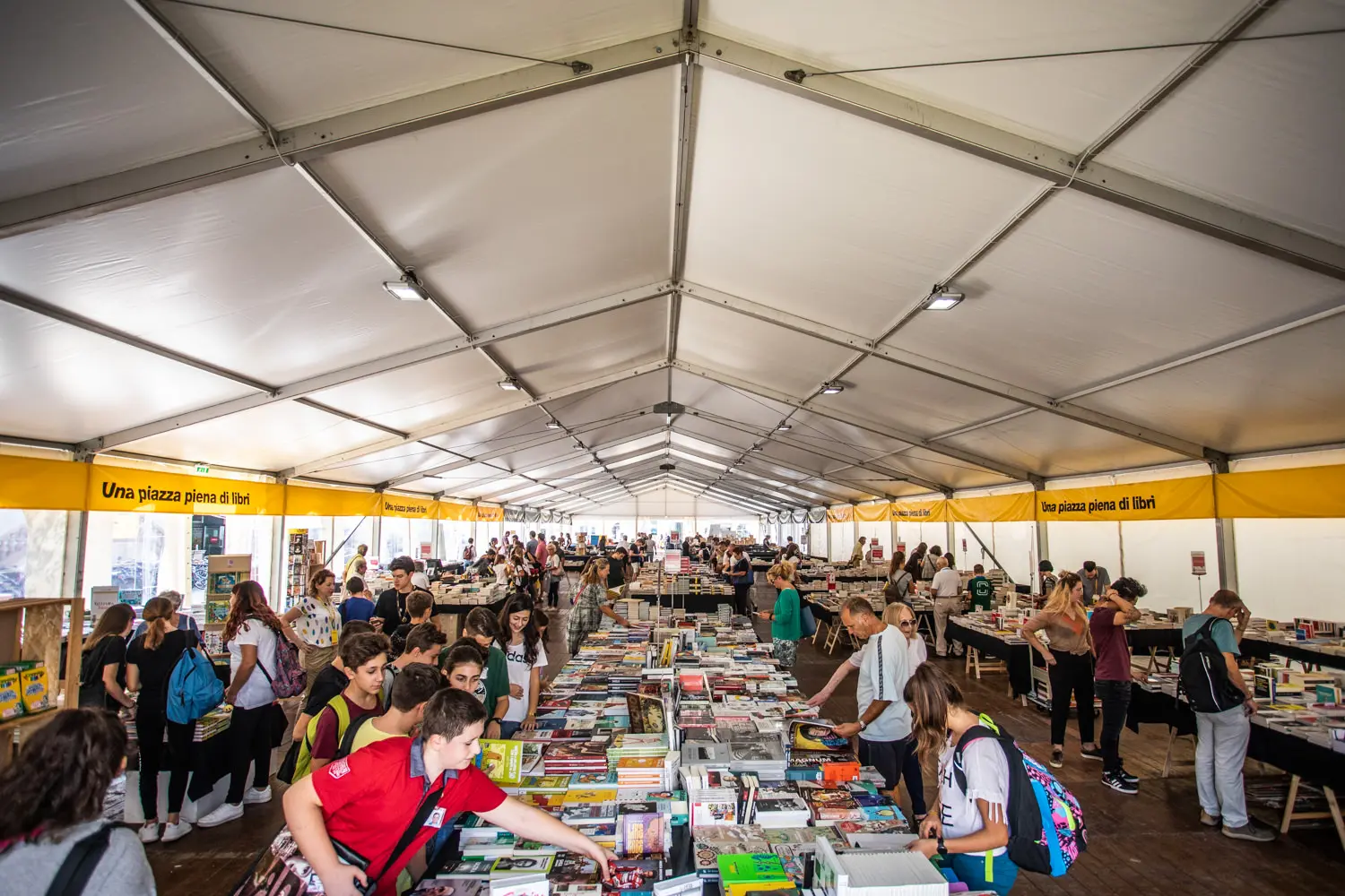 Le persone sfogliano i libri su lunghi tavoli all'interno di una grande tenda bianca. Lo spazio è pieno di attività mentre individui di varie età esplorano e raccolgono libri. Sono visibili striscioni gialli e l'atmosfera è vivace e coinvolgente.
