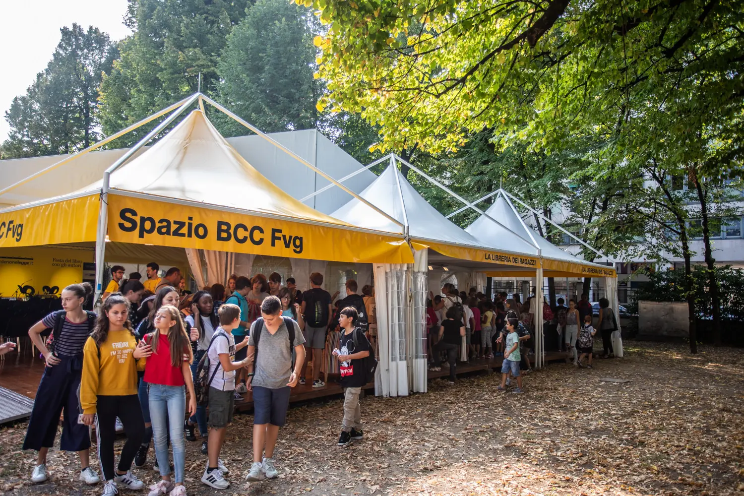 Un gruppo di persone, tra cui bambini e adulti, si trova all'esterno di una serie di tende gialle e bianche con la scritta "Spazio BCC Fvg". L'ambientazione è un'area esterna con alberi e foglie cadute a terra, a suggerire un raduno o un evento.