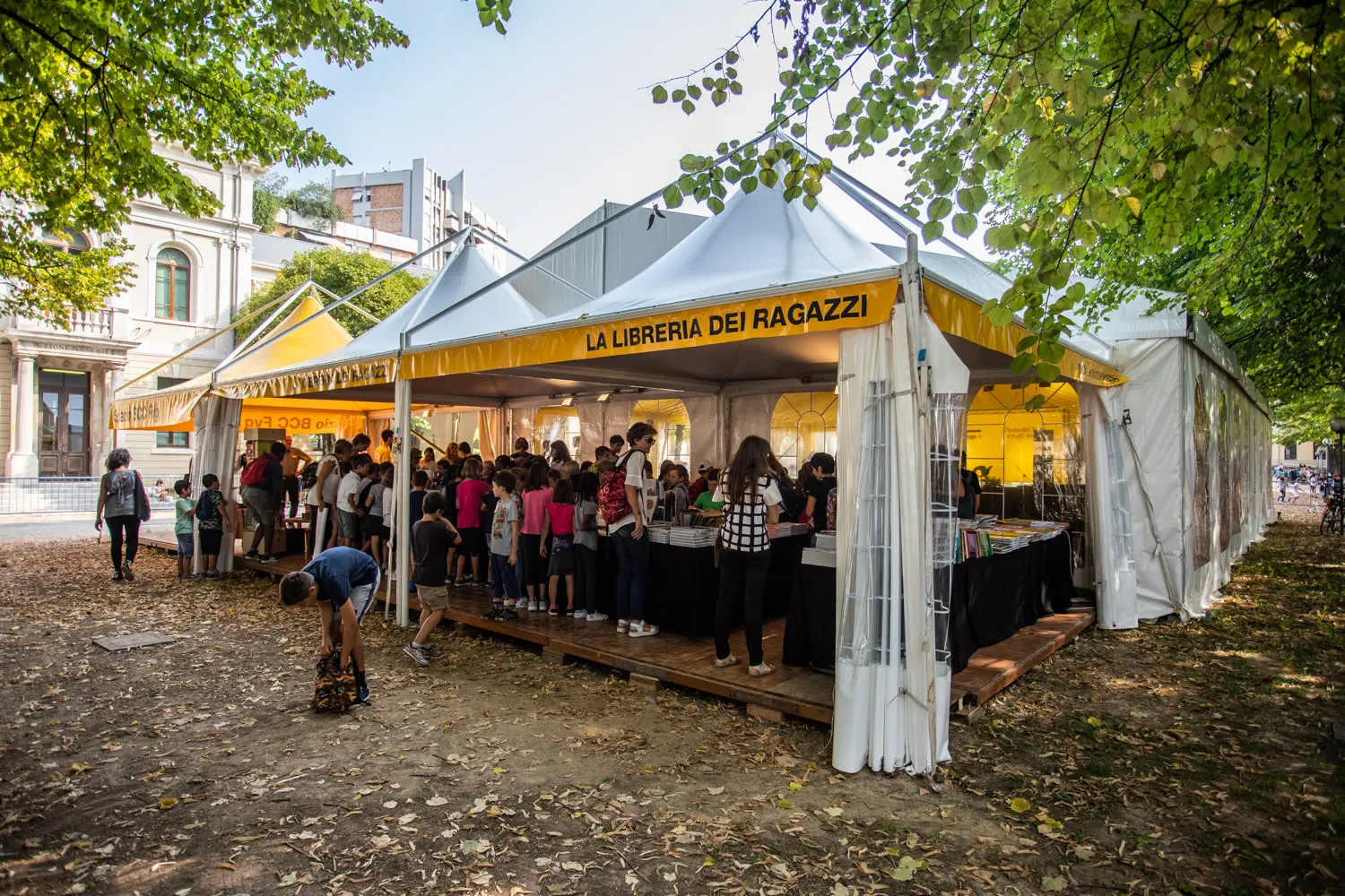 Fiera del libro all'aperto sotto una grande tenda bianca con uno striscione giallo con la scritta "La Libreria Dei Ragazzi". Le persone sfogliano i libri all'interno, mentre altri sono in fila. Alberi con foglie verdi circondano l'area e foglie cadute ricoprono il terreno.