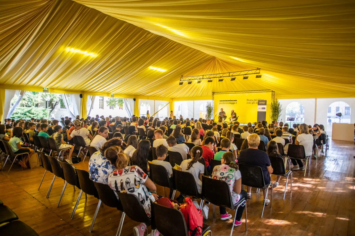 Un folto gruppo di persone sedute su sedie sotto una spaziosa tenda gialla, attente di fronte a un palco con relatori. L'evento si svolge durante il giorno, con luce naturale che filtra attraverso la tenda.