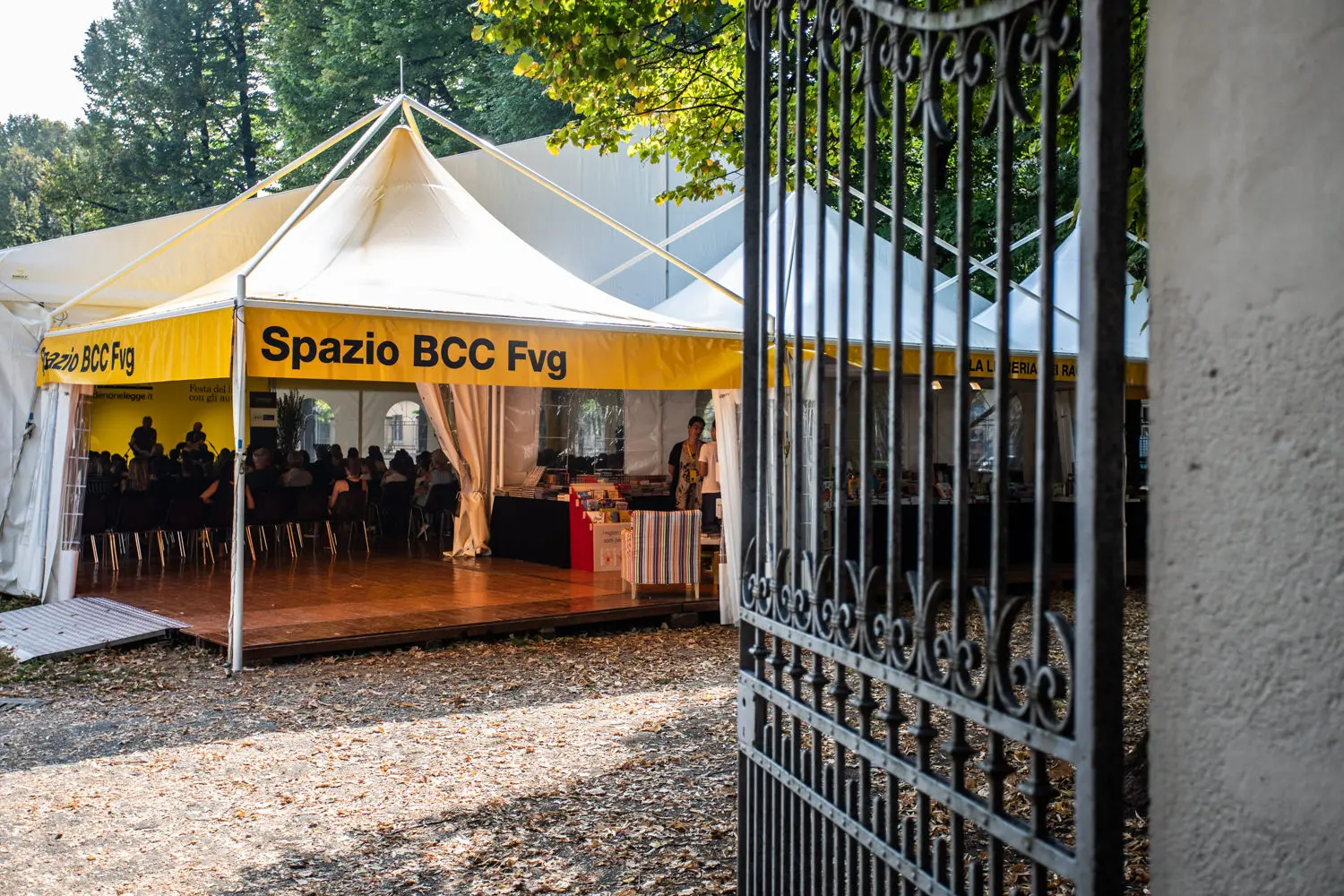 All'esterno è allestita una tenda gialla e bianca con la scritta "Spazio BCC Fvg" sul lato. Le persone sono sedute all'interno, visibili attraverso le pareti trasparenti della tenda. La scena è incorniciata da un cancello aperto in ferro battuto e circondata da alberi.