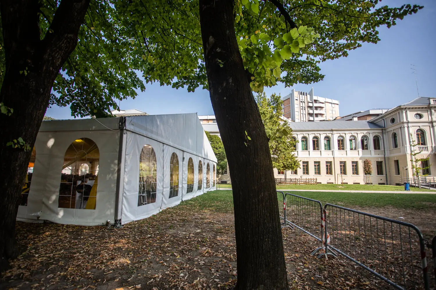 Una grande tenda bianca con finestre ad arco è installata su un'area erbosa accanto a un edificio storico. Alberi con foglie verdi oscurano parzialmente la vista. Barricate metalliche sono posizionate sulla destra e sullo sfondo sono visibili edifici moderni.