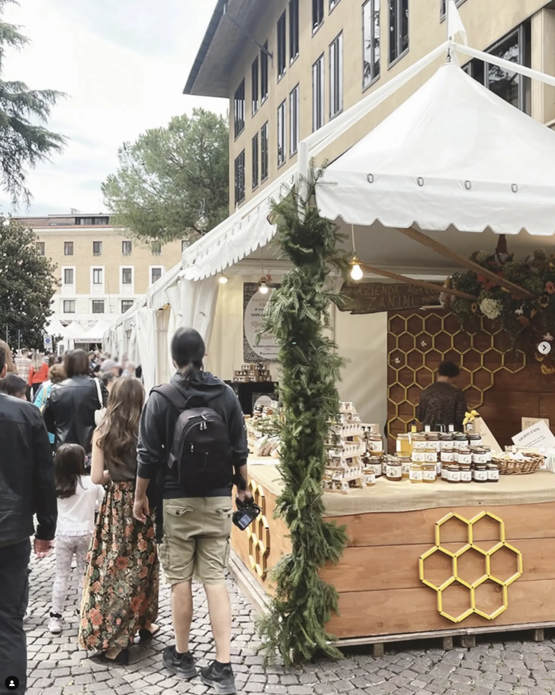 La gente cammina lungo un sentiero acciottolato in un mercato all'aperto. Una tenda bianca espone prodotti a base di miele, con decorazioni esagonali e ghirlande verdi. Alberi e un grande edificio sono sullo sfondo.