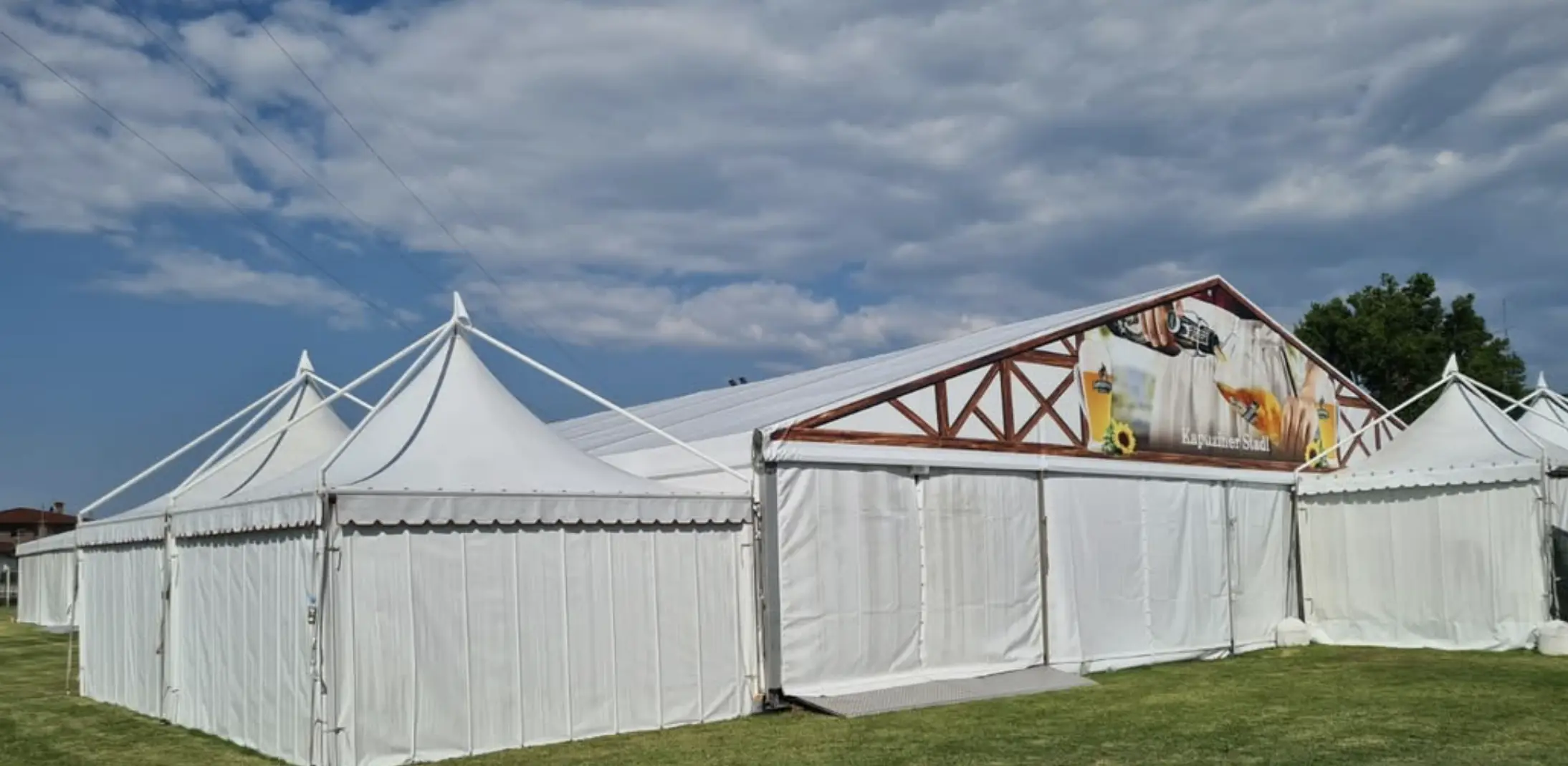 Una grande tenda bianca con più tetti a punta e una struttura in legno è adagiata su un'area erbosa sotto un cielo parzialmente nuvoloso. Uno striscione colorato decora la parte anteriore della tenda.