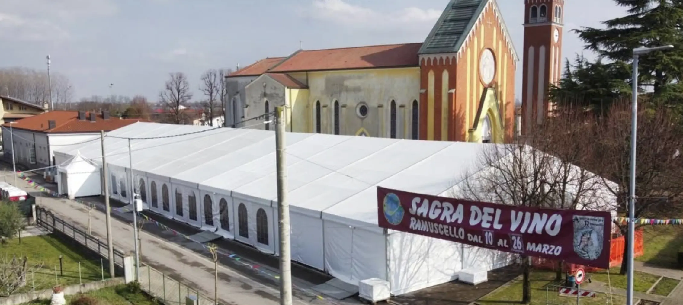 Grandi tende bianche sono state installate di fronte a un edificio storico e colorato della chiesa. Uno striscione in italiano indica una festa del vino dall'11 al 26 marzo. Alberi spogli e un cielo limpido sono sullo sfondo.