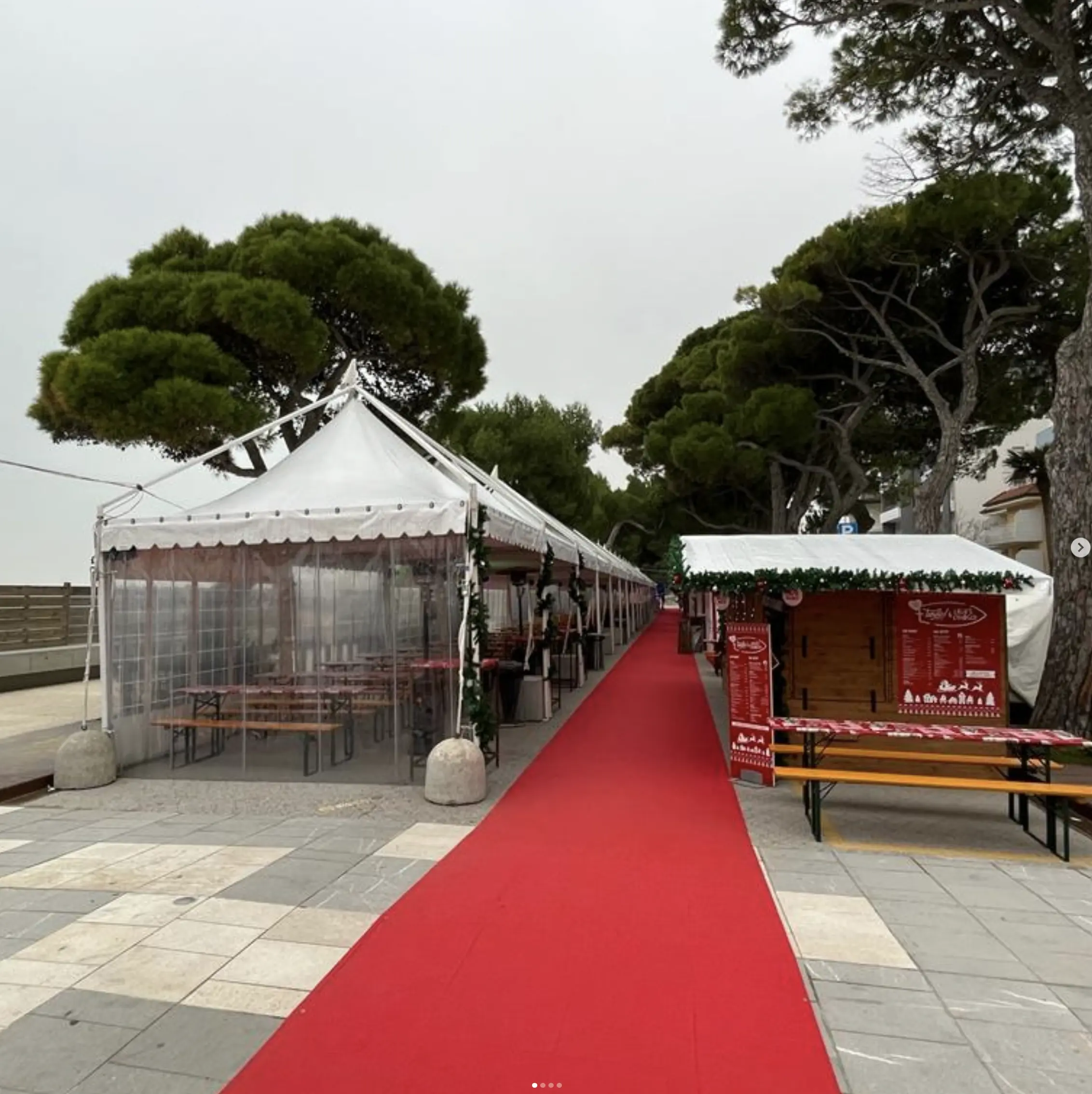 Un lungo tappeto rosso attraversa una fila di tende festive all'aperto e bancarelle di legno, allestite sotto grandi alberi. L'area sembra preparata per un evento, con panchine sotto le tende e luci a corda visibili. Il cielo è coperto.