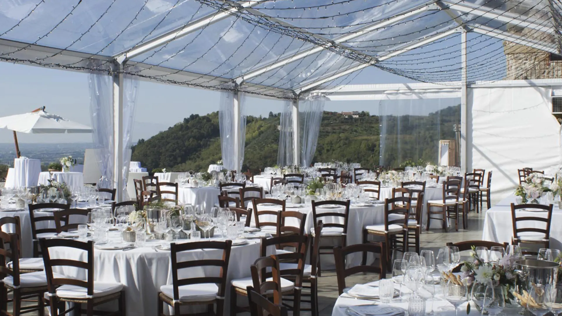 Una location per matrimoni all'aperto splendidamente allestita sotto una tenda trasparente. Tavoli rotondi coperti da tovaglie bianche e circondati da sedie in legno scuro. Ogni tavolo è decorato con centrotavola floreali, bicchieri e apparecchiatoi. Le colline panoramiche sono visibili all'esterno.