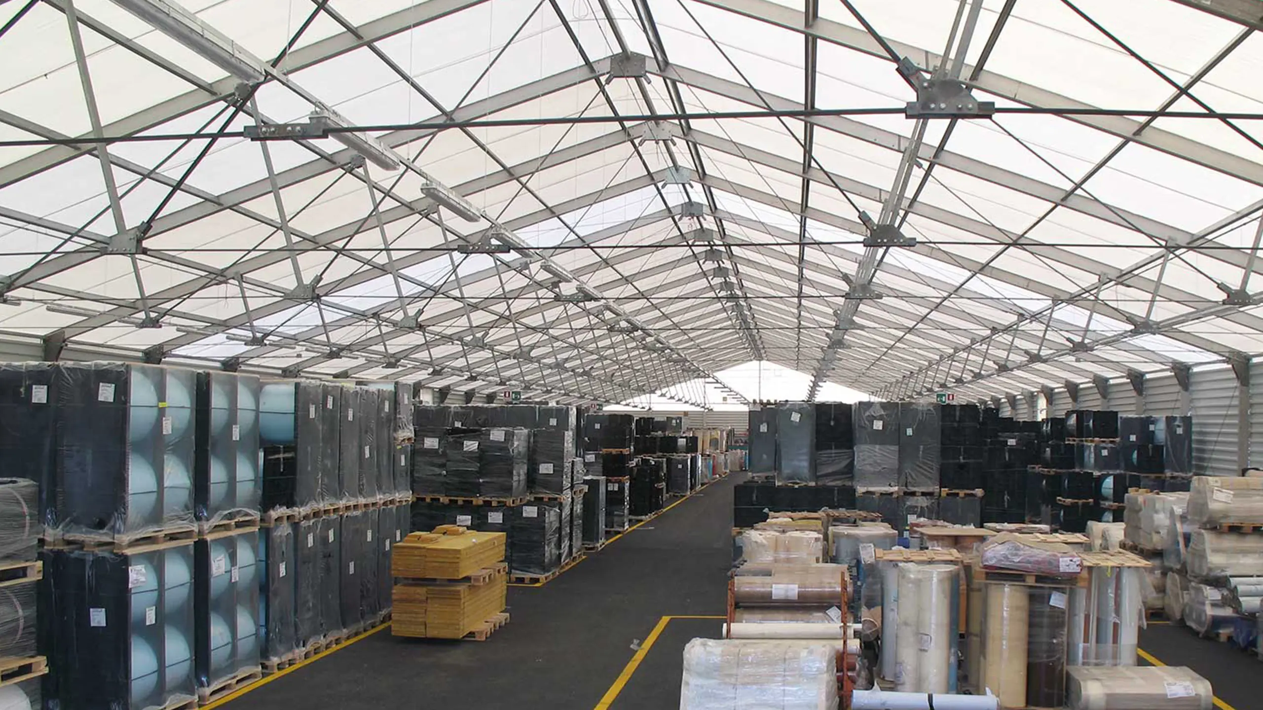 Vista interna di un grande magazzino con soffitto alto, che espone file di pallet impilati contenenti vari beni e materiali. Il magazzino ha una struttura in metallo ed è inondato di luce naturale dai pannelli traslucidi del tetto.