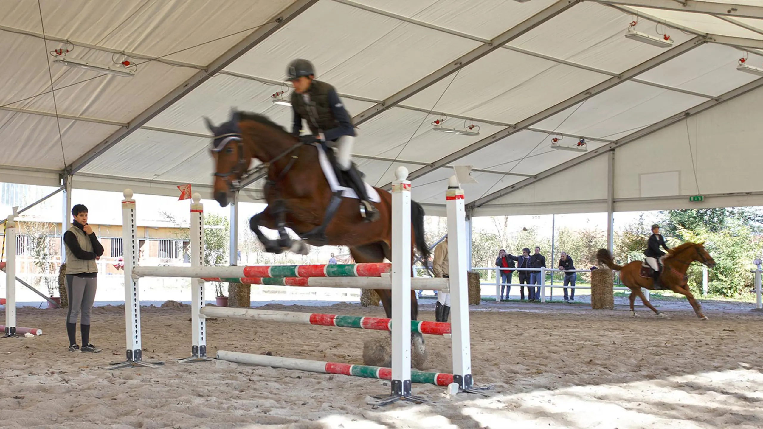 Una persona cavalca un cavallo, saltando un ostacolo in un'arena equestre al coperto. Un altro cavallo e cavaliere sono visibili sullo sfondo, con alcuni spettatori in piedi nelle vicinanze. L'arena è coperta da un grande tetto.