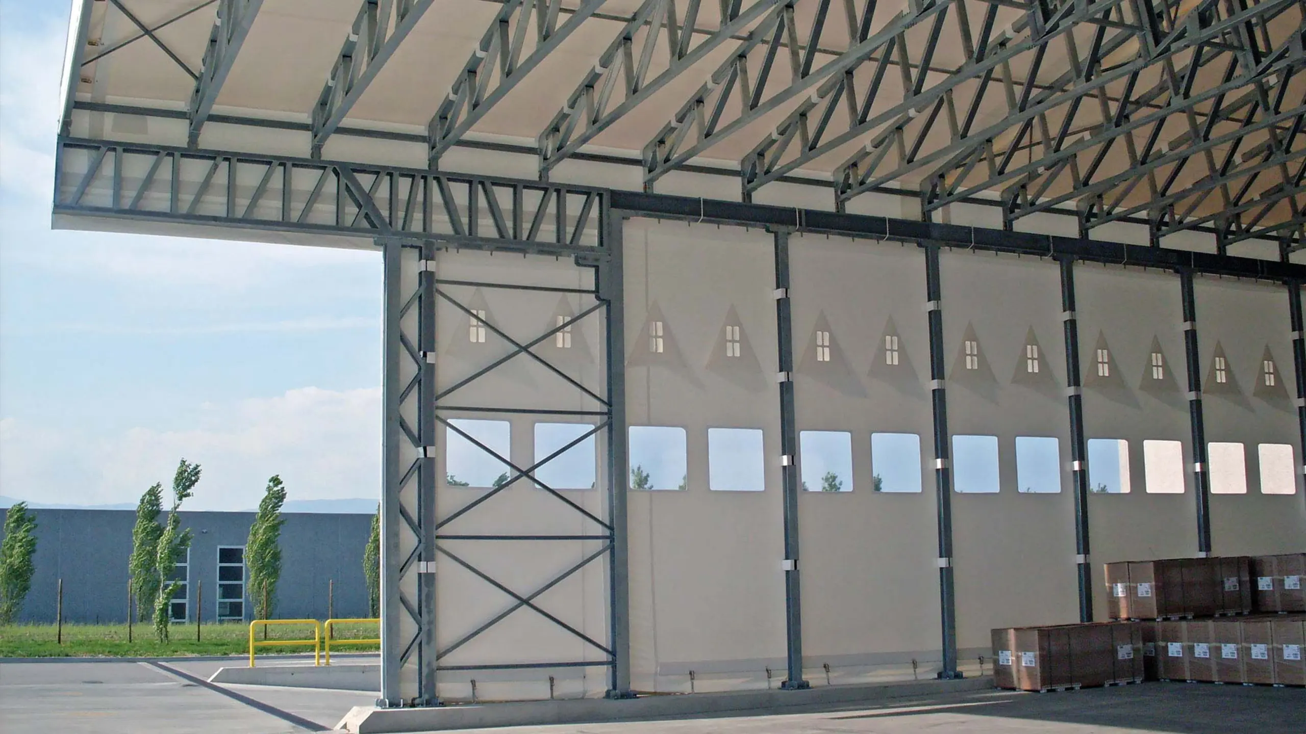 Vista interna di un magazzino industriale con una grande porta scorrevole aperta. La struttura presenta travi metalliche e un soffitto alto. Ci sono scatole di cartone accatastate sulla destra e l'esterno mostra un'area pavimentata e un cielo parzialmente nuvoloso.