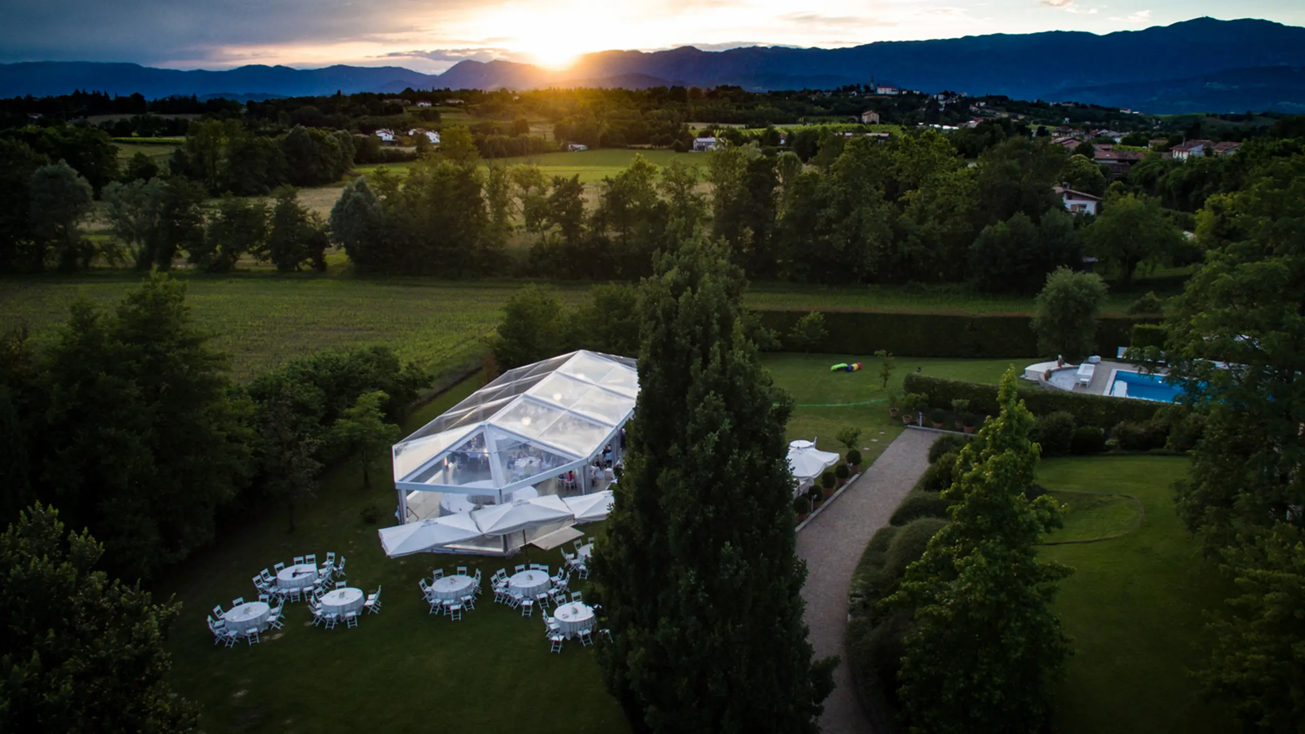Una vista panoramica al tramonto mostra una tenda trasparente allestita su un prato verde lussureggiante con diversi tavoli rotondi sistemati all'esterno. Gli alberi circostanti incorniciano l'area, con un sentiero che conduce verso la tenda. Le montagne sono visibili sullo sfondo.