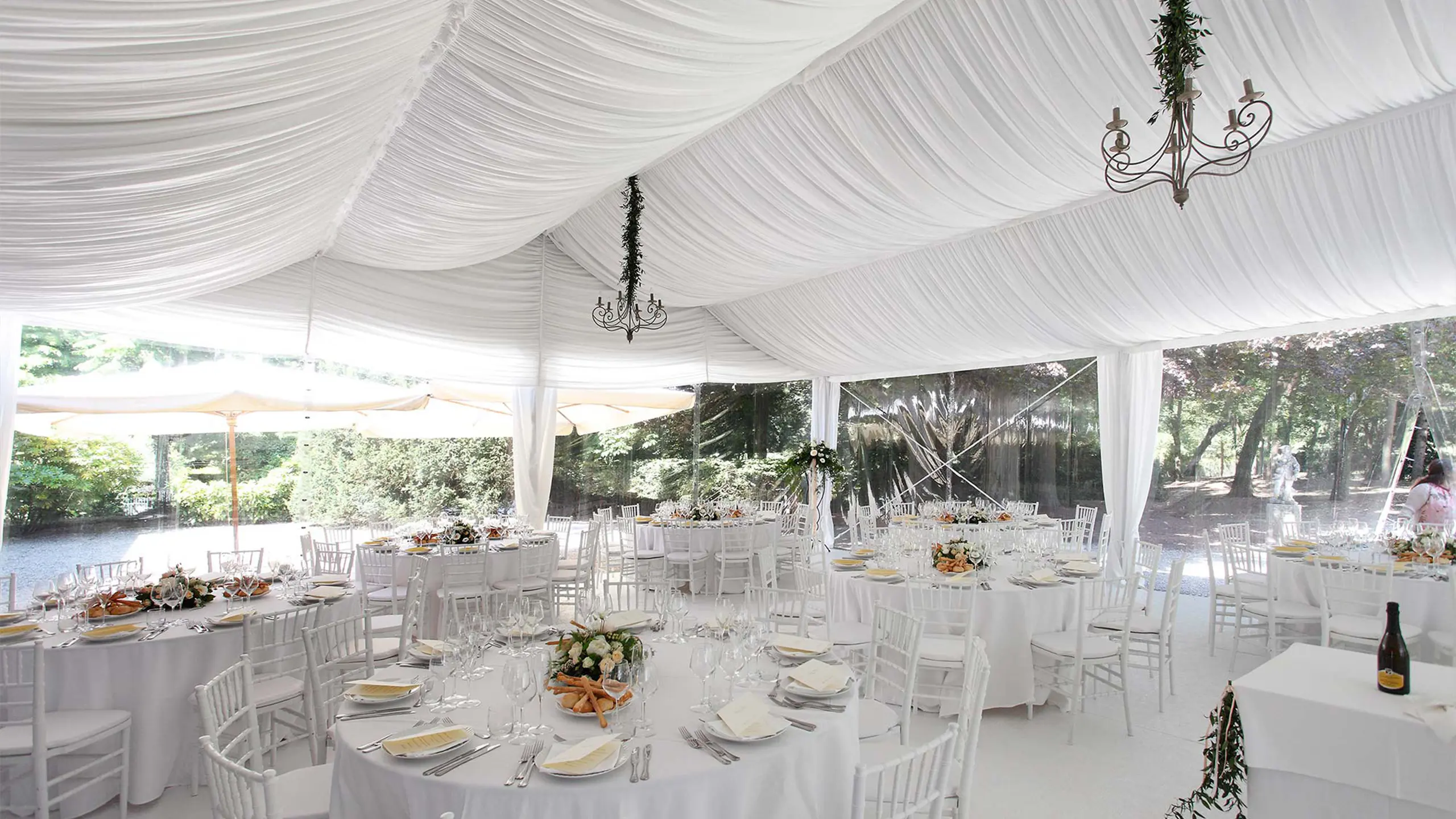 Una tenda bianca con eleganti drappeggi ospita un allestimento formale per eventi. I tavoli rotondi sono adornati con tovaglie bianche, centrotavola floreali e apparecchiati ordinatamente. La luce naturale filtra attraverso i lati trasparenti e i lampadari pendono dal soffitto.