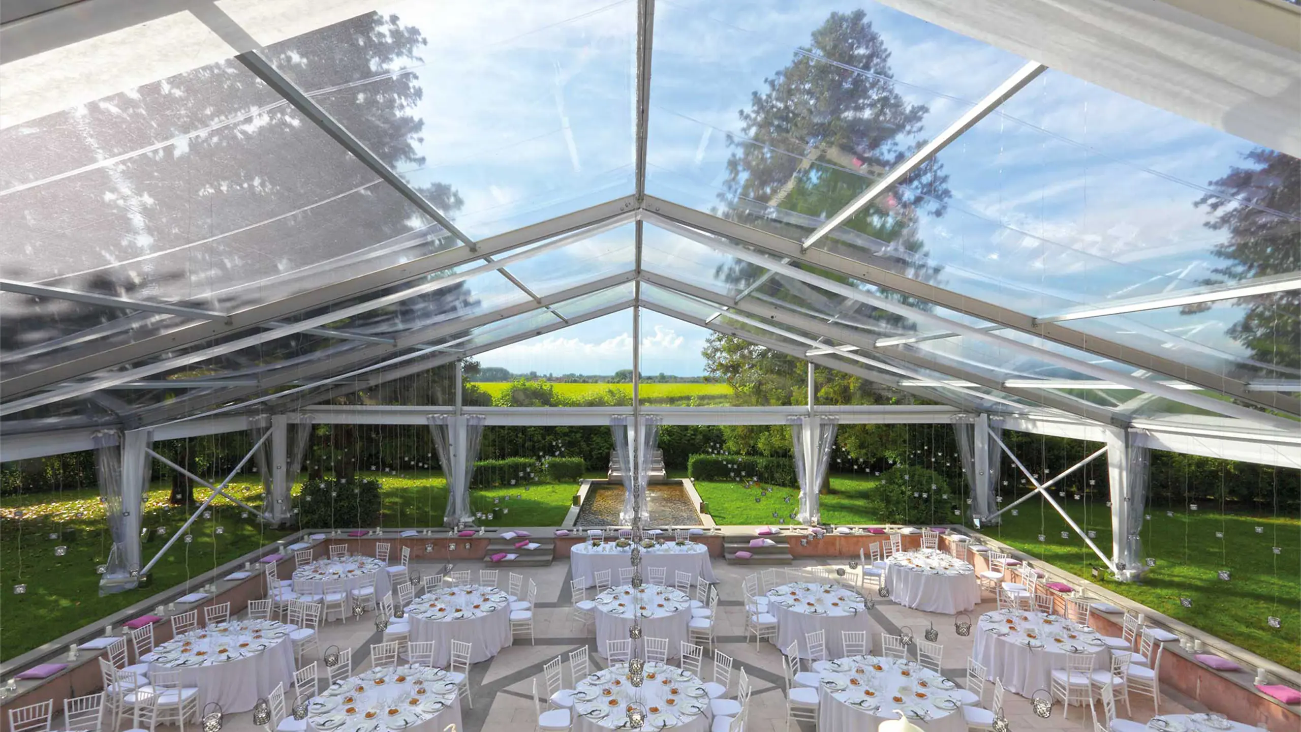 Un'ampia location per matrimoni all'aperto sotto una tenda trasparente, con tavoli rotondi con tovaglie e sedie bianche. Un prato verde lussureggiante e alberi circondano la struttura, con uno sfondo di un ampio campo aperto sotto un cielo azzurro.
