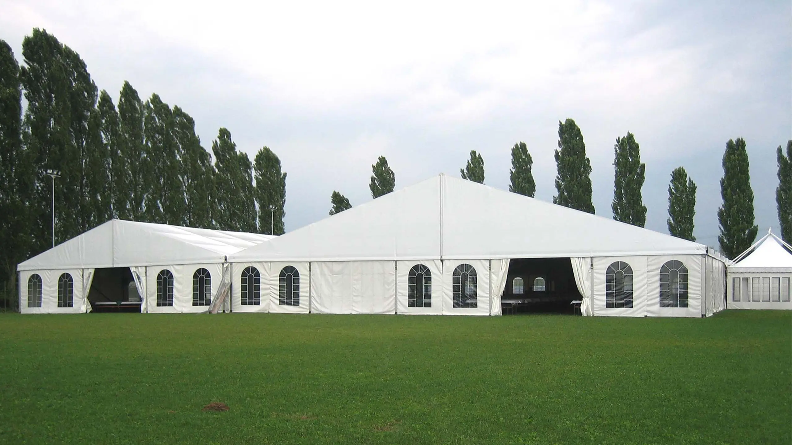 Un grande tendone bianco per eventi allestito su un'area erbosa, circondato da alberi alti. Il tendone ha finestre ad arco e un tetto a punta, e il cielo è nuvoloso sullo sfondo.