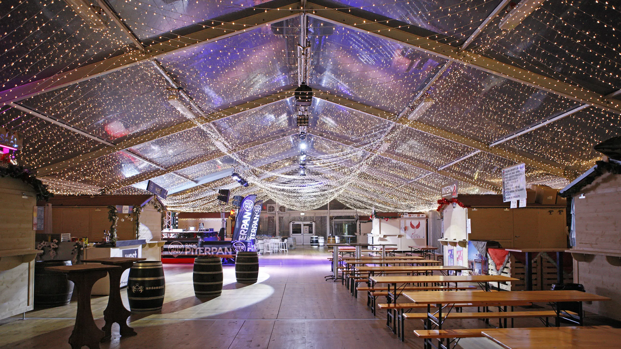 Interno di una tenda festiva con tavoli e panche in legno, decorata con luci a corda sul soffitto. Botti e piccoli stand di cibo fiancheggiano i lati e sono visibili cartelli, creando un'atmosfera accogliente.