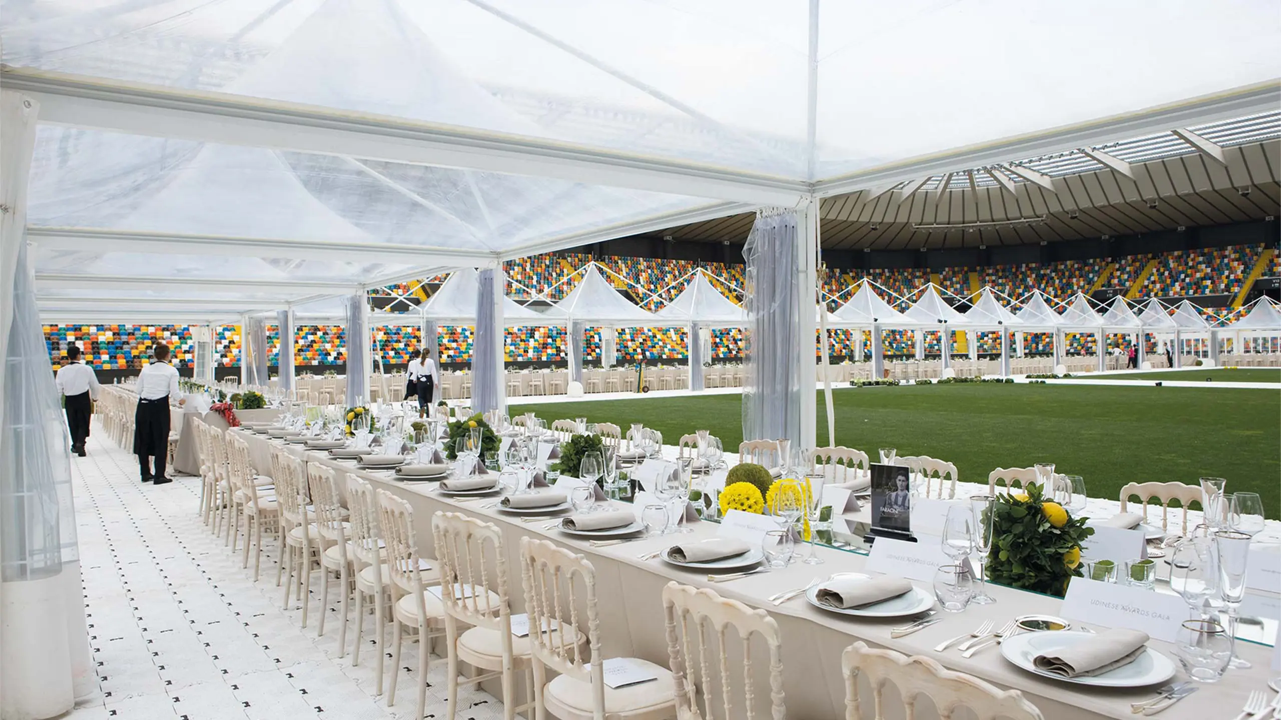 Un lungo tavolo da pranzo elegantemente apparecchiato sotto una tenda trasparente in uno stadio. Il tavolo è decorato con piatti bianchi, posate, fiori gialli e verde. Camerieri in camice bianco preparano per gli ospiti. File di tende bianche circondano il campo verde.
