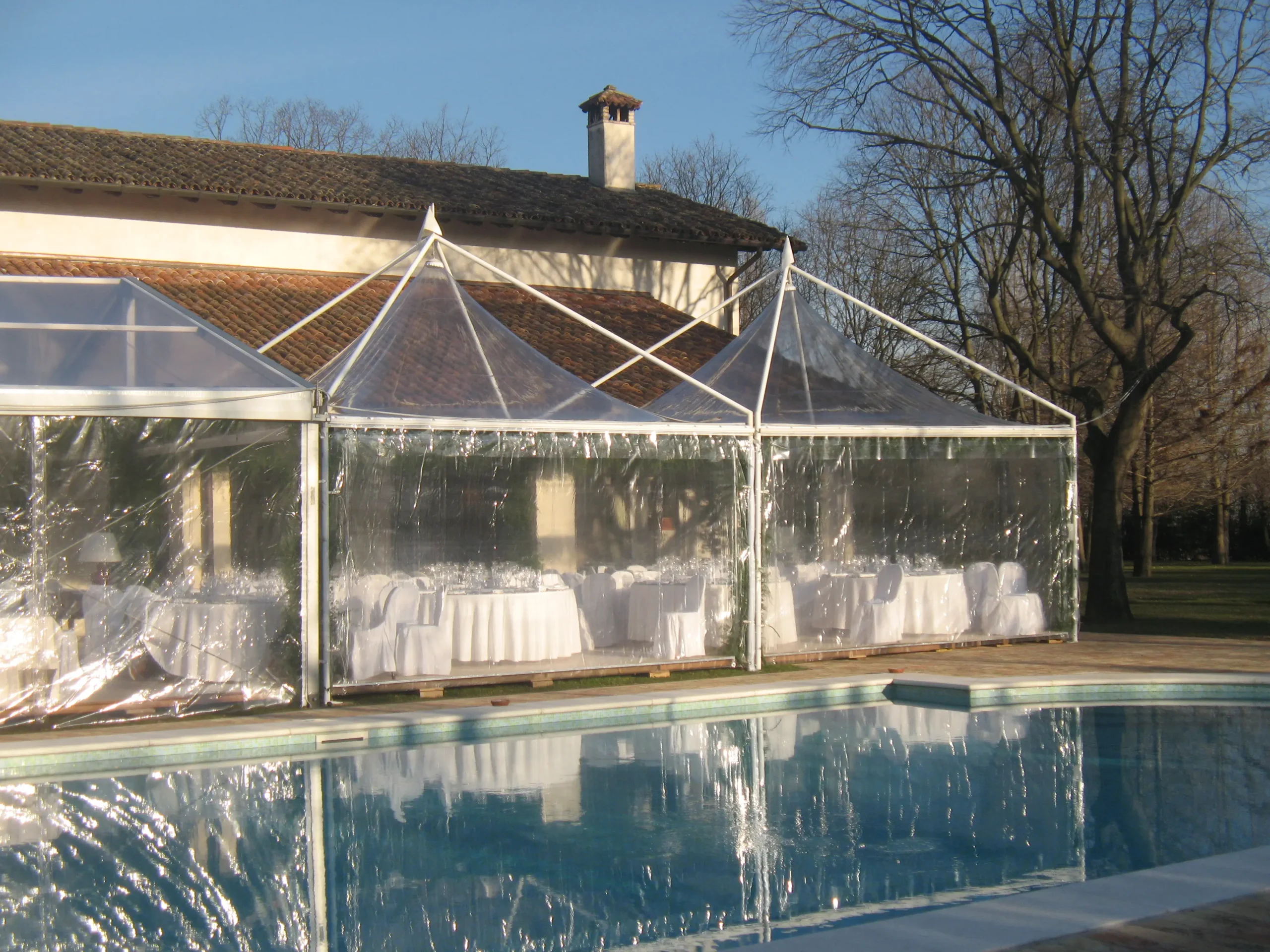 Una scena serena all'aperto con un tendone trasparente allestito accanto a una piscina. All'interno del tendone ci sono tavoli elegantemente apparecchiati con tovaglie e sedie bianche, che suggeriscono un evento formale. Lo sfondo include un edificio rustico e alberi spogli.