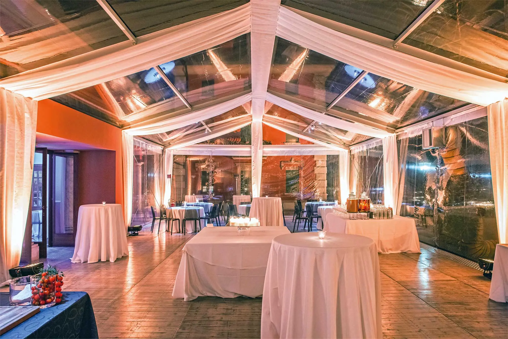 Una tenda trasparente con tessuto drappeggiato ospita tavoli ricoperti di stoffa bianca. Ci sono tavoli rotondi e rettangolari, alcuni con piccole candele. La tenda è ben illuminata, creando un'atmosfera calda.
