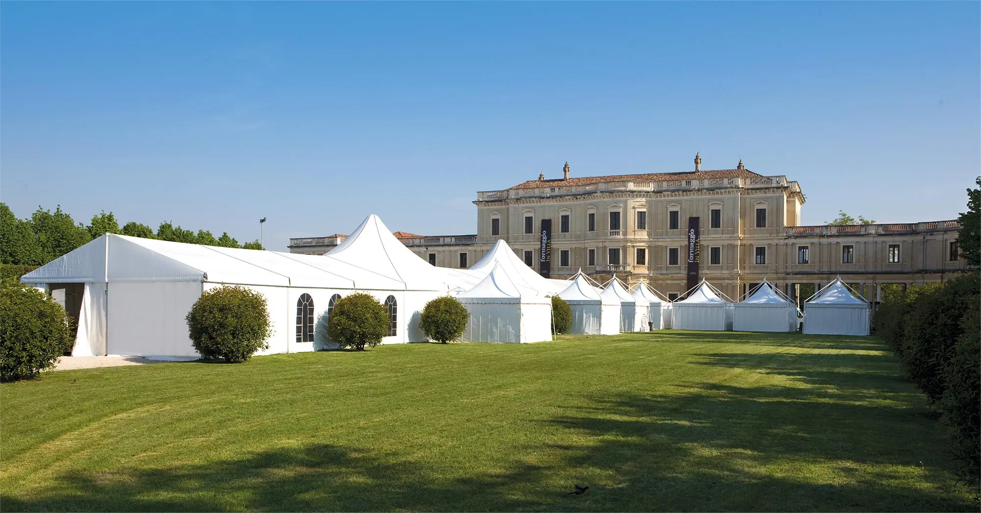 Una serie di tende bianche sono state installate su un grande prato ben curato di fronte a un edificio storico. Il cielo è limpido e azzurro e i cespugli sono stati sistemati attorno alle tende, aggiungendo un tocco di scenografia all'ambiente esterno.