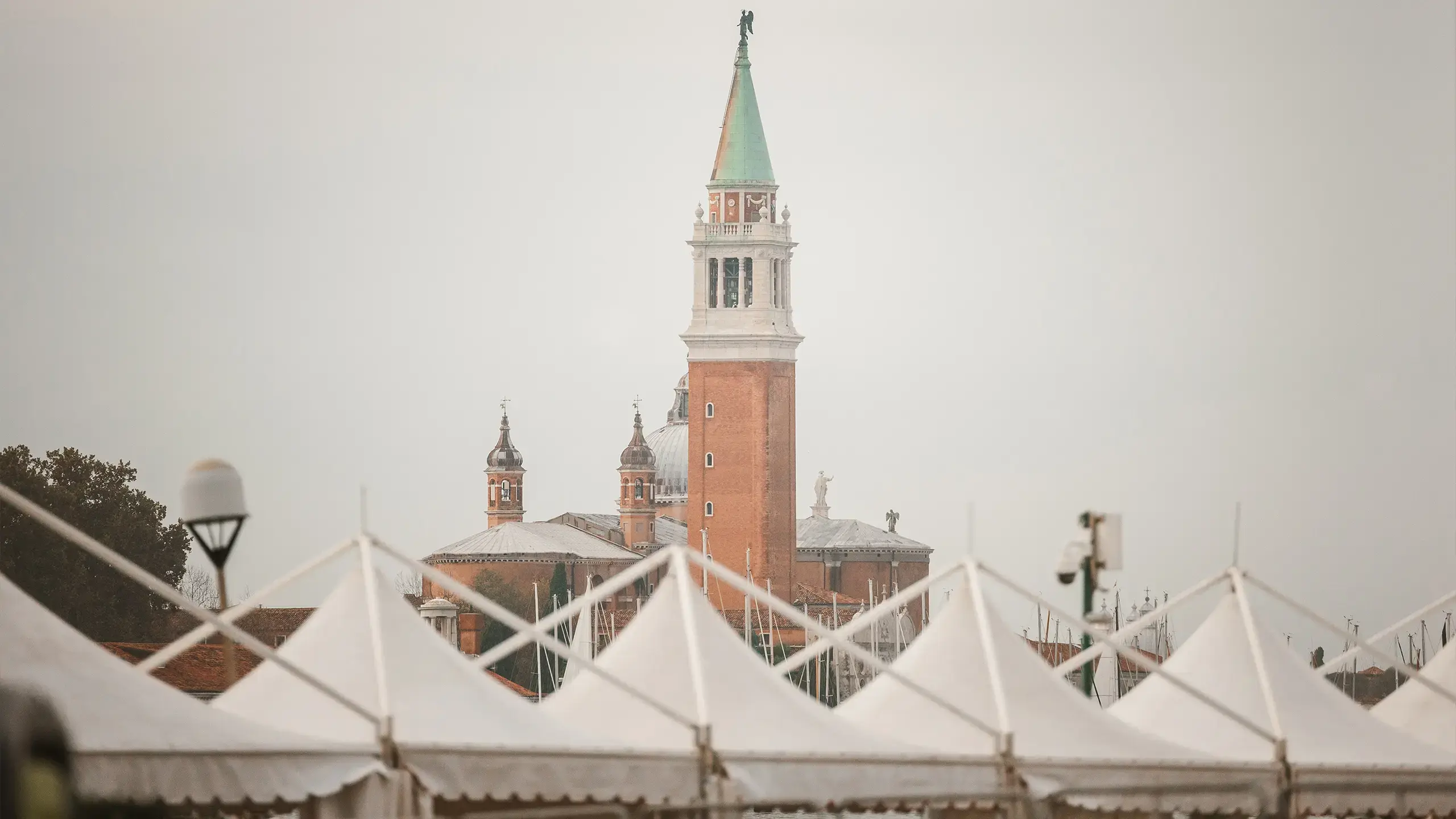 L'immagine mostra un'alta torre in mattoni rossi con una guglia verde, che ricorda il Campanile di San Giorgio Maggiore a Venezia. In primo piano ci sono tende bianche con la punta, che oscurano parzialmente la vista della torre. Il cielo è coperto.