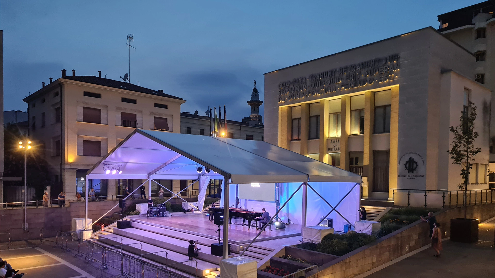 Un grande palco all'aperto con un baldacchino bianco è allestito di fronte a un edificio illuminato al tramonto. Un pianoforte a coda nero è sul palco e alcune persone sono visibili nelle vicinanze. Il cielo si sta oscurando, con alcune nuvole presenti.