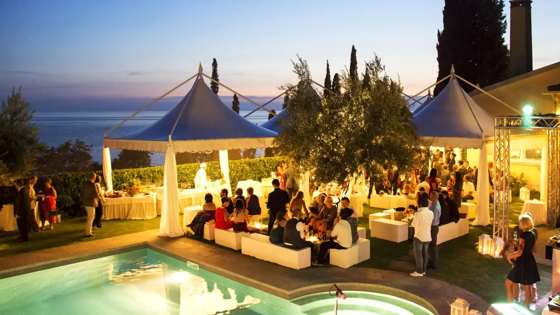 Un evento serale all'aperto a bordo piscina con persone che socializzano sotto tende a baldacchino bianco. L'ambientazione include alberi e si affaccia sull'oceano al tramonto. Gli ospiti sono seduti su mobili bianchi, creando un'atmosfera vivace ed elegante.