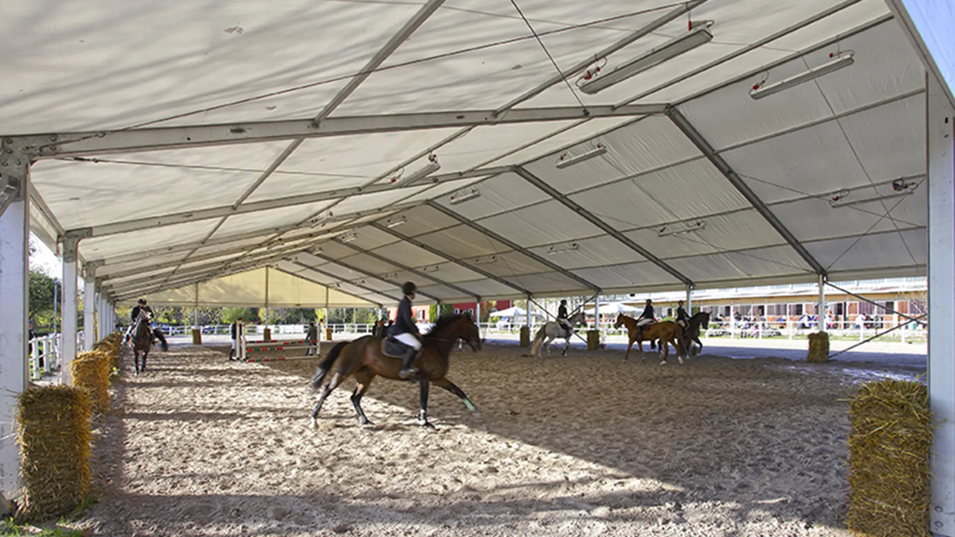 Arena equestre coperta con più cavalieri a cavallo che si esercitano. Lo spazio è coperto da una grande tenda bianca e circondato da balle di fieno. La luce naturale entra da un lato.