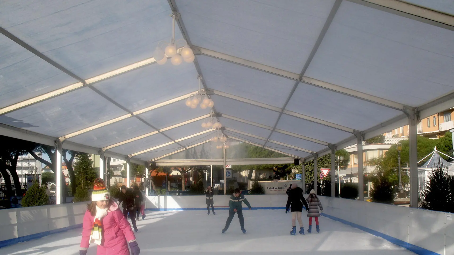 Le persone pattinano sul ghiaccio su una pista all'aperto coperta da una grande tenda bianca. La scena include bambini e adulti, alcuni in movimento e altri in piedi. Alberi ed edifici sono visibili sullo sfondo. Il tempo è limpido e soleggiato.