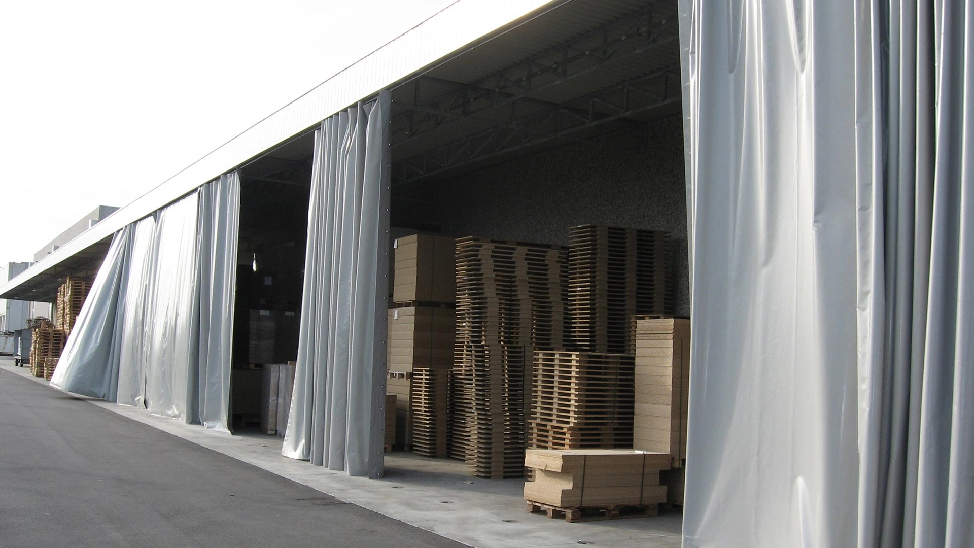 Un magazzino con baie aperte, parzialmente coperte da grandi teloni grigi. All'interno, sono visibili pile di pallet di legno. La scena è su una superficie di cemento con un tetto di un edificio che si estende sull'area.