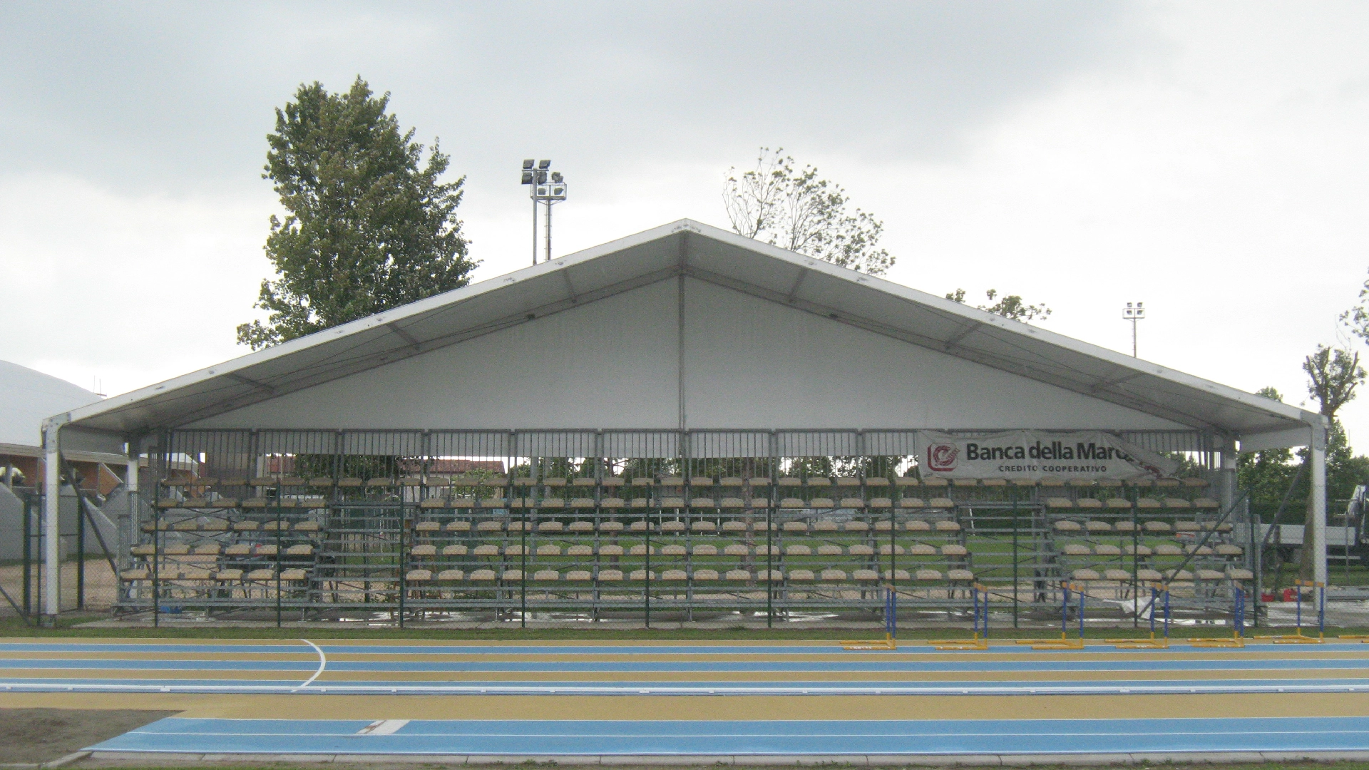 Tribuna esterna sotto un tetto bianco di riparo in un campo sportivo. Le tribune hanno una struttura metallica verde e posti a sedere a due livelli. Un cartello recita "Banca della Marca". In primo piano c'è una pista da corsa con corsie blu e gialle. Cielo coperto sopra.
