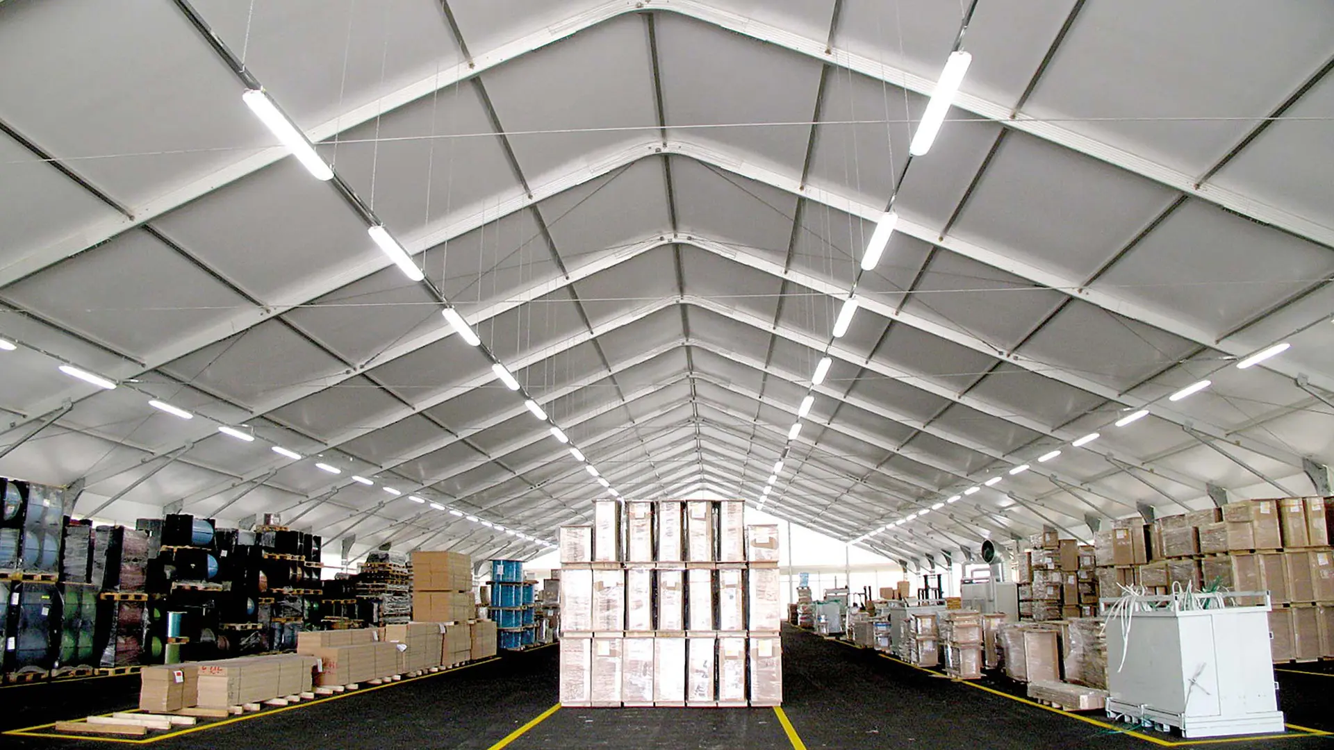 Vista interna di un grande magazzino con soffitti alti, con pile di scatole e pallet disposti ordinatamente su entrambi i lati. Il corridoio centrale è libero e luci fluorescenti in alto illuminano lo spazio.