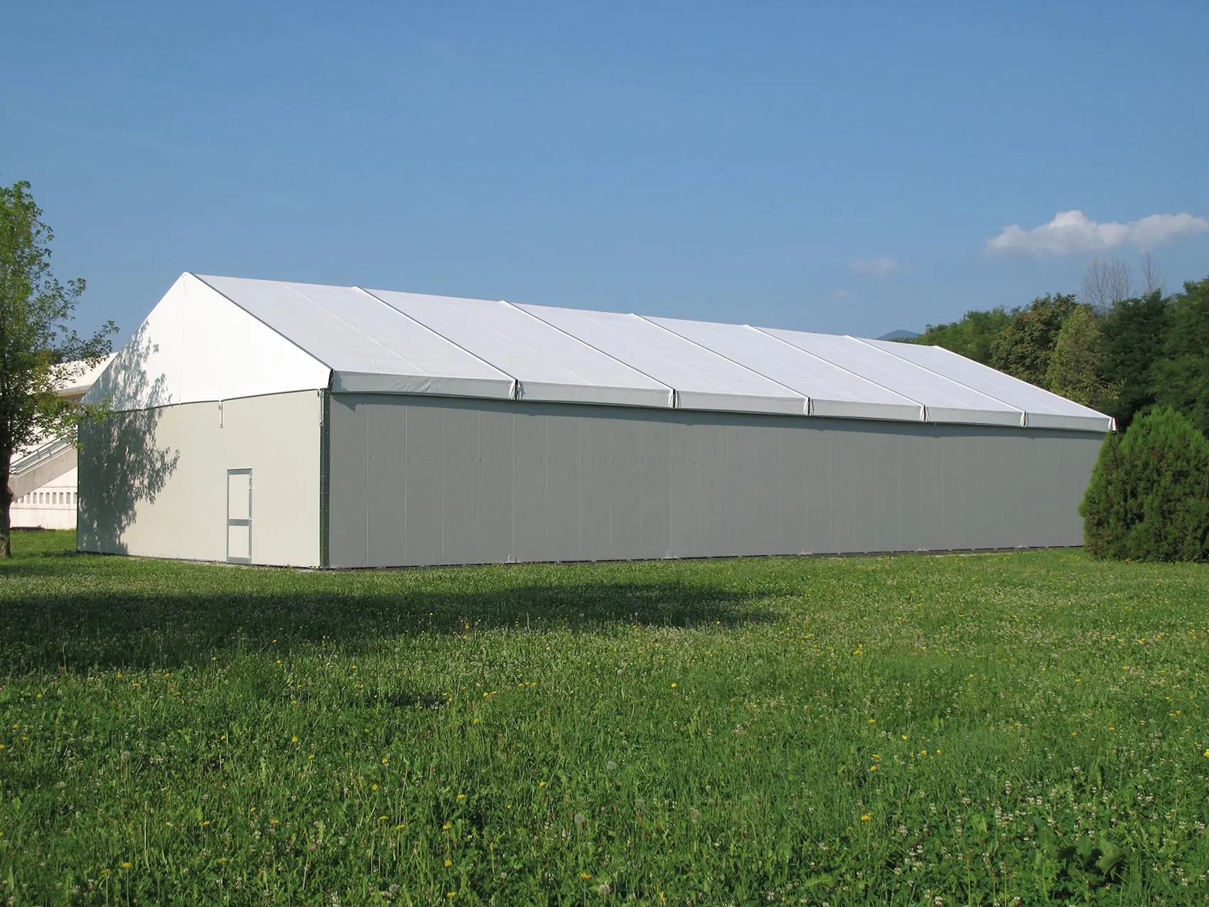 Una grande tenda bianca rettangolare con un tetto a punta si erge su un campo erboso sotto un cielo azzurro terso. Piccoli alberi e cespugli sono visibili sullo sfondo.