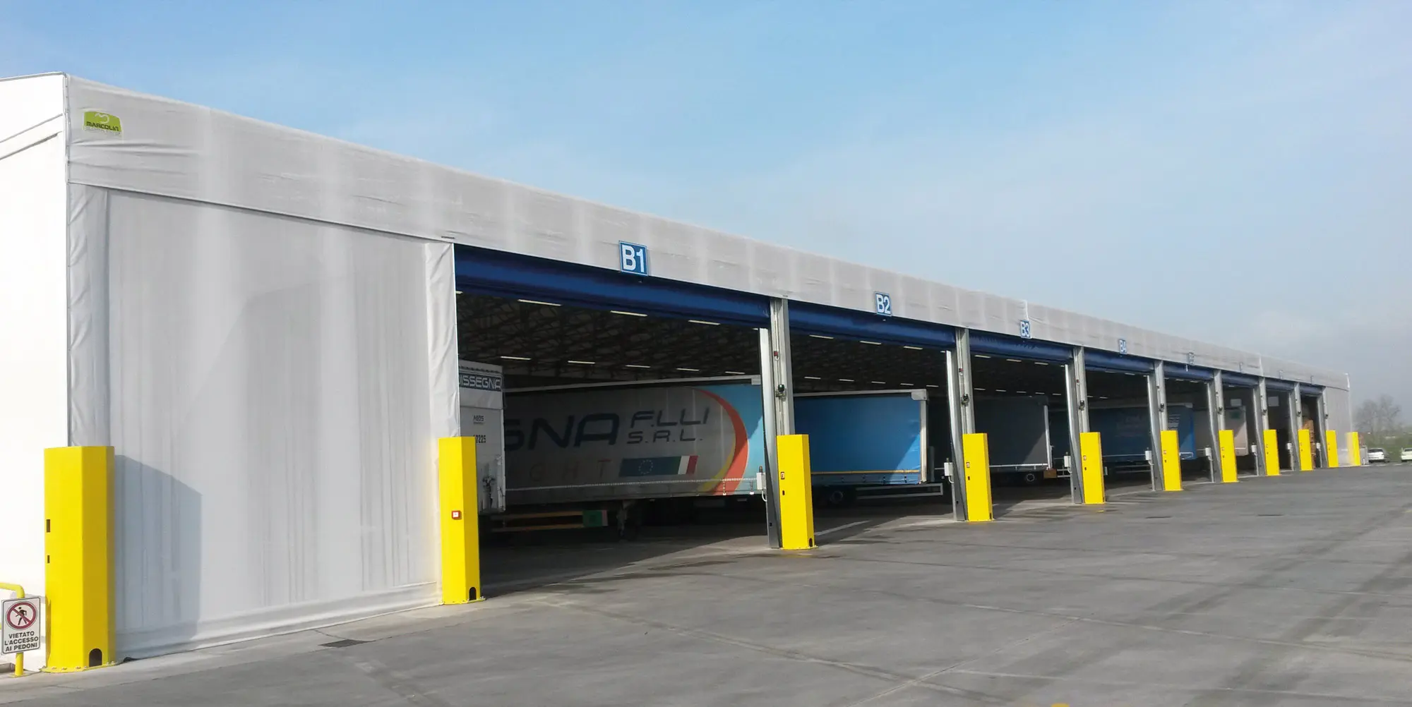 Una fila di banchine di carico con rimorchi per camion appoggiati alle baie. Le banchine sono coperte da una grande struttura a baldacchino e ogni baia ha un numero blu sopra. Bitte di colore giallo brillante delimitano l'area di fronte alle banchine.