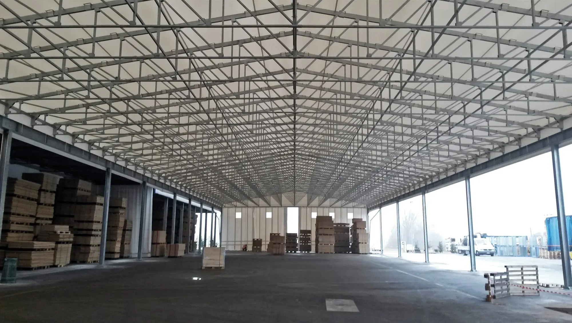 Interno di un grande magazzino con un soffitto alto con struttura metallica. Lungo le pareti e al centro sono disposte diverse pile di scatole di cartone e pallet. Lo spazio è ben illuminato dalla luce naturale proveniente dalle grandi finestre.