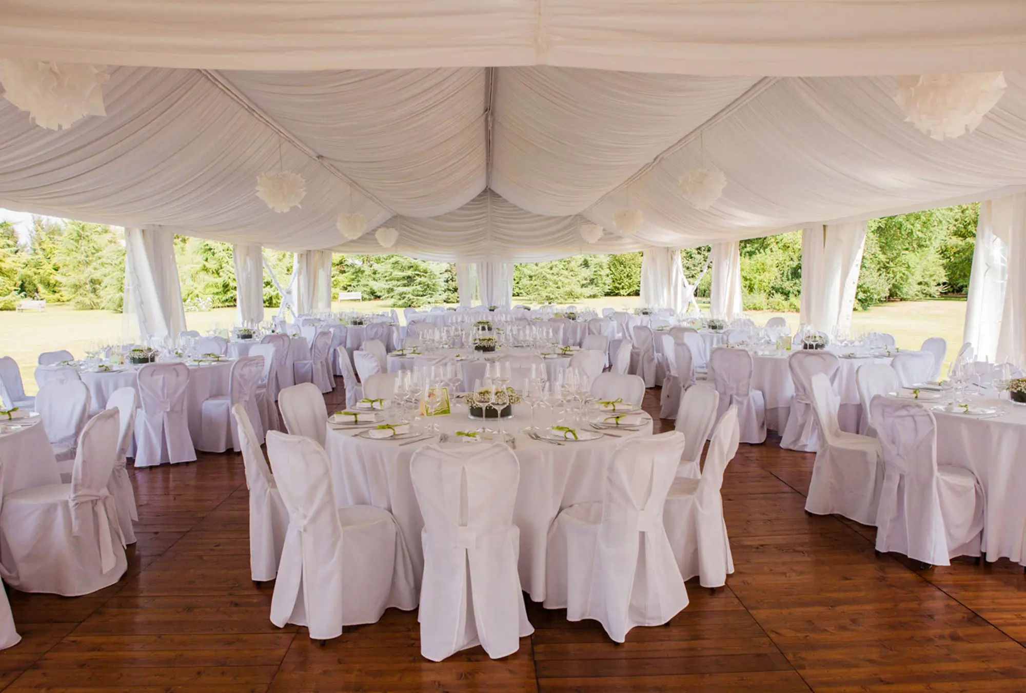 Una spaziosa tenda bianca con pavimento in legno è allestita per un evento. I tavoli rotondi sono elegantemente disposti con tovaglie bianche e decorati con bicchieri e centrotavola. Sedie bianche circondano ogni tavolo e la tenda si apre su una vista di alberi verdi.