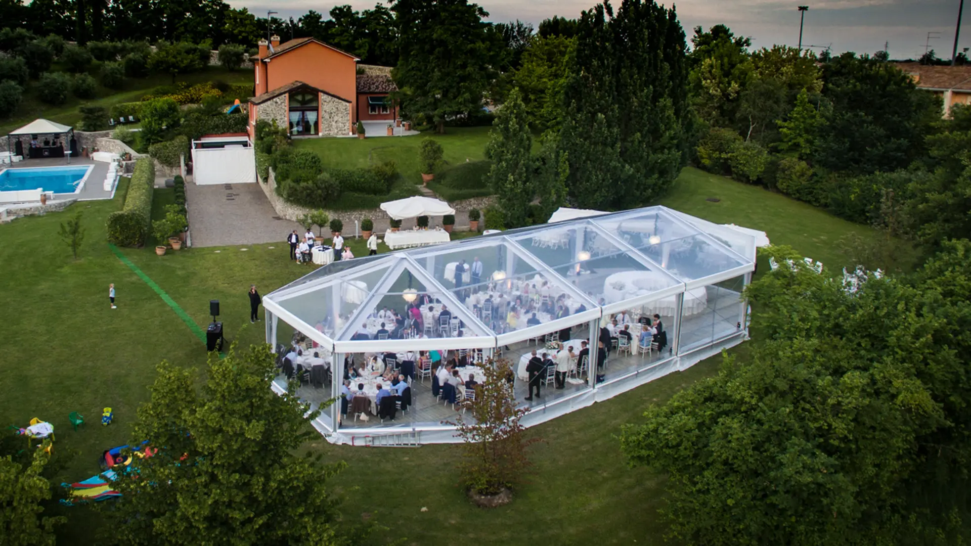 Vista aerea di un ricevimento di nozze in una tenda trasparente su un prato verde lussureggiante. La location è circondata da alberi, con una casa rossa e una piscina visibili sullo sfondo. Gli ospiti sono seduti all'interno della tenda.
