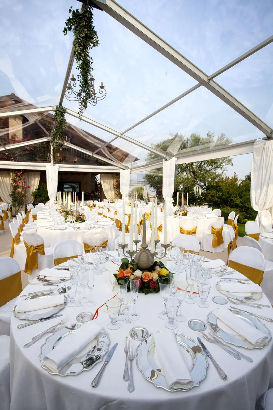 Un tavolo per il ricevimento nuziale splendidamente apparecchiato sotto una tenda trasparente. I tavoli sono adornati con tovaglie bianche, sedie con accenti dorati, eleganti posate e centrotavola floreali. Lo sfondo è caratterizzato da una vegetazione lussureggiante e un cielo azzurro terso.