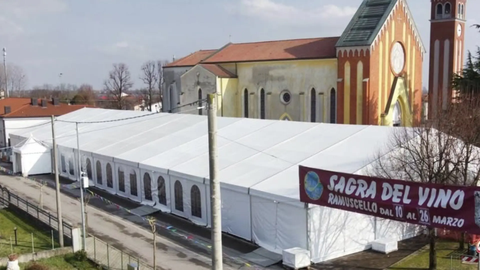 Un grande tendone bianco per eventi è stato allestito vicino a un edificio storico della chiesa. Uno striscione sul tendone recita "Sagra del Vino, Ramusello dal 10 al 26 Marzo". La zona è tranquilla, con alcuni alberi e un palo della luce nelle vicinanze.