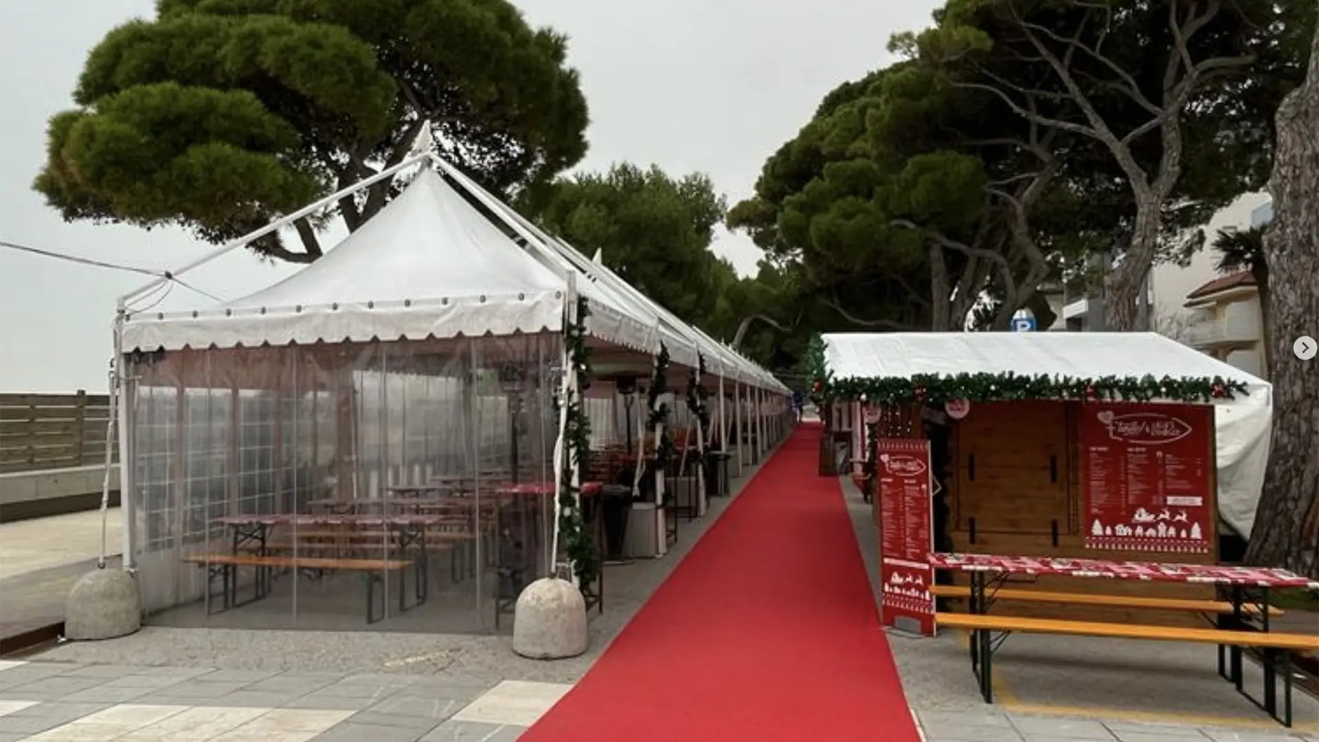 Bancarelle di mercatini all'aperto sotto tende bianche con tende trasparenti, allineate lungo un tappeto rosso. Tavoli e panche di legno sono visibili all'interno delle tende. Alberi alti con fitte chiome verdi sono sullo sfondo. Cielo coperto sopra.